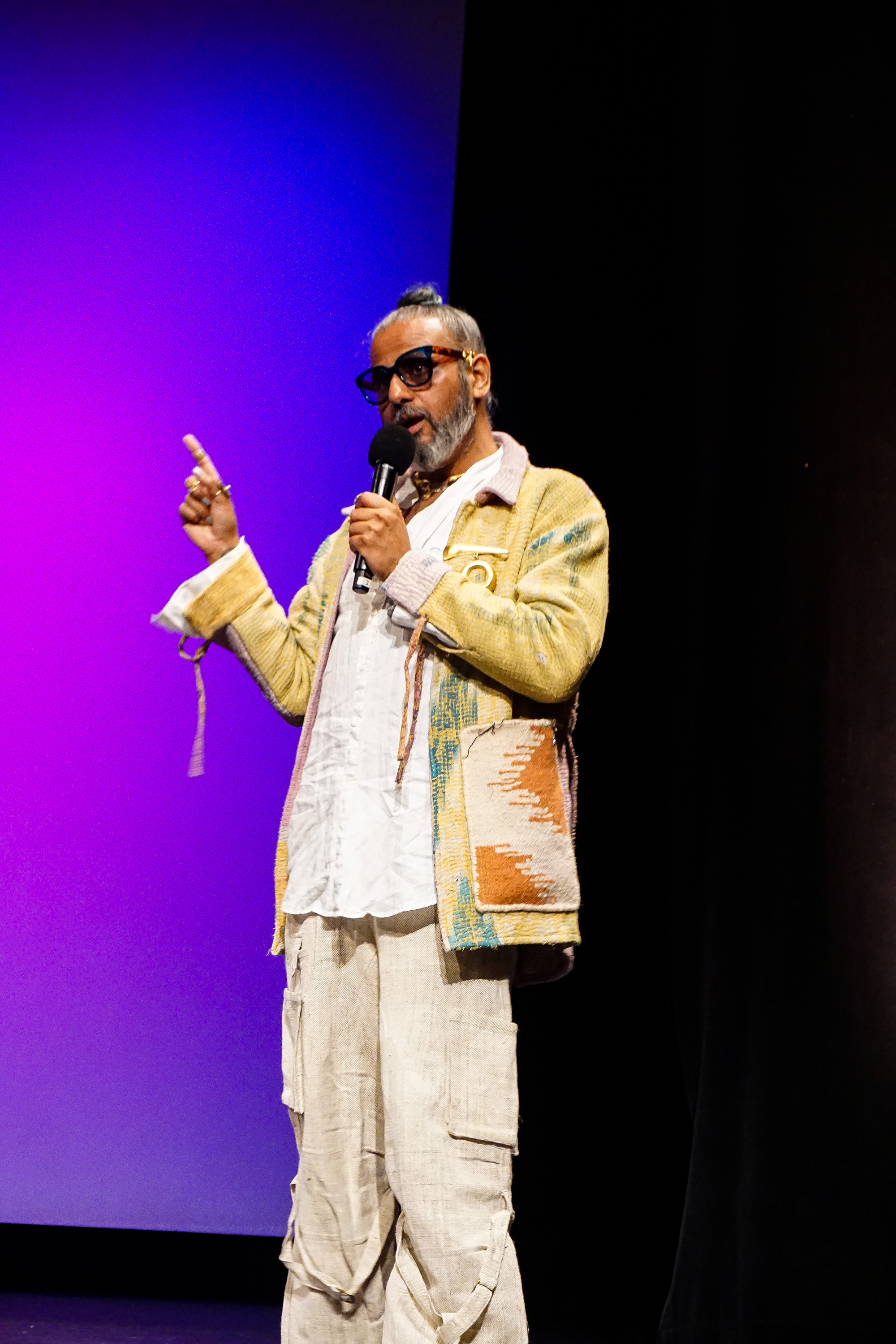 Ajay Shrivastav standing on stage speaking into a microphone and point up while wearing a yellow jacket, sunglasses and golden jewelry