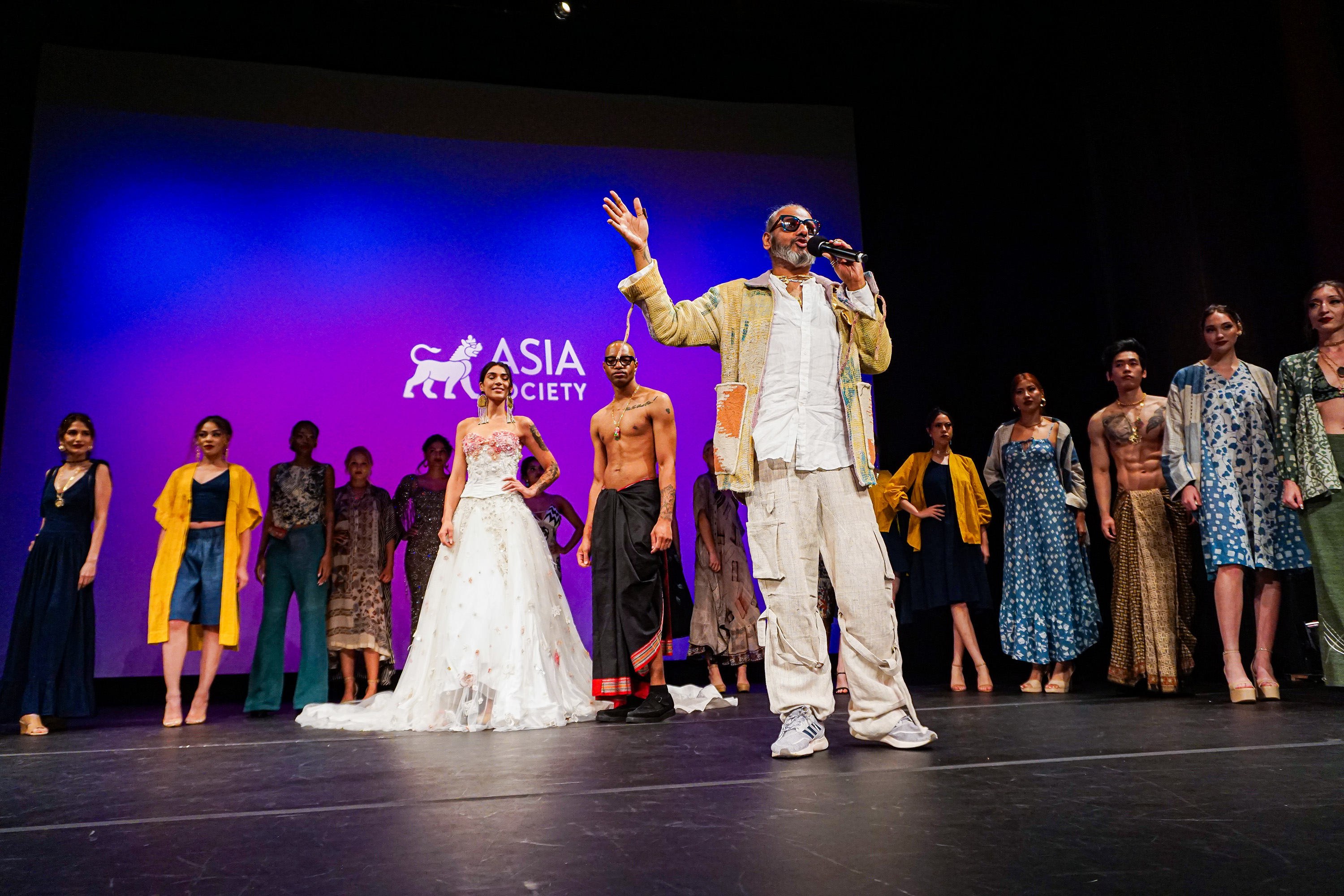 Ajay Shrivastav gesticulating to the crowd and speaking in the microphone in front of crowd of models on the runway