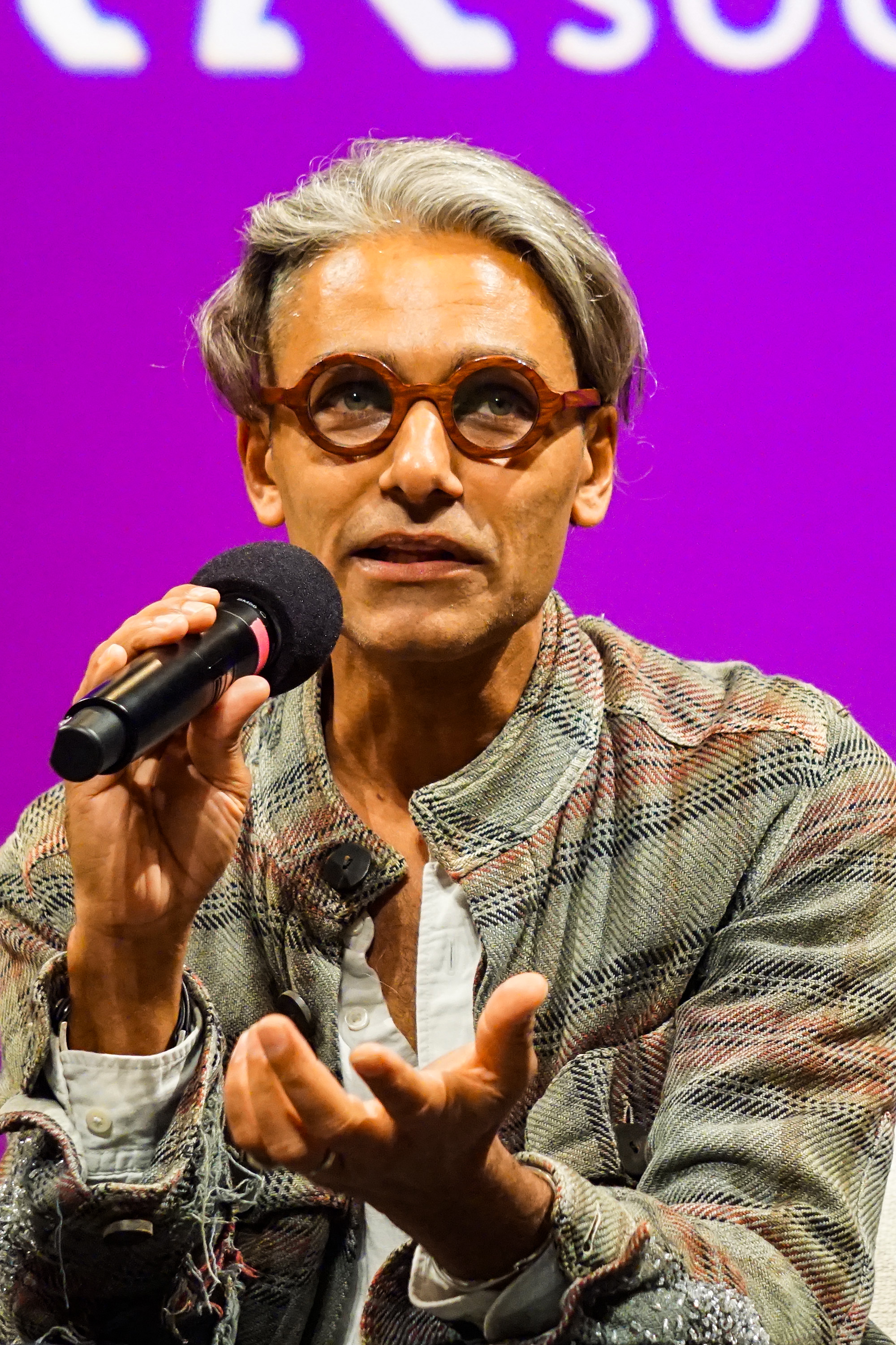 Sandy Dalal speaking into a microphone with his hand out to the viewer