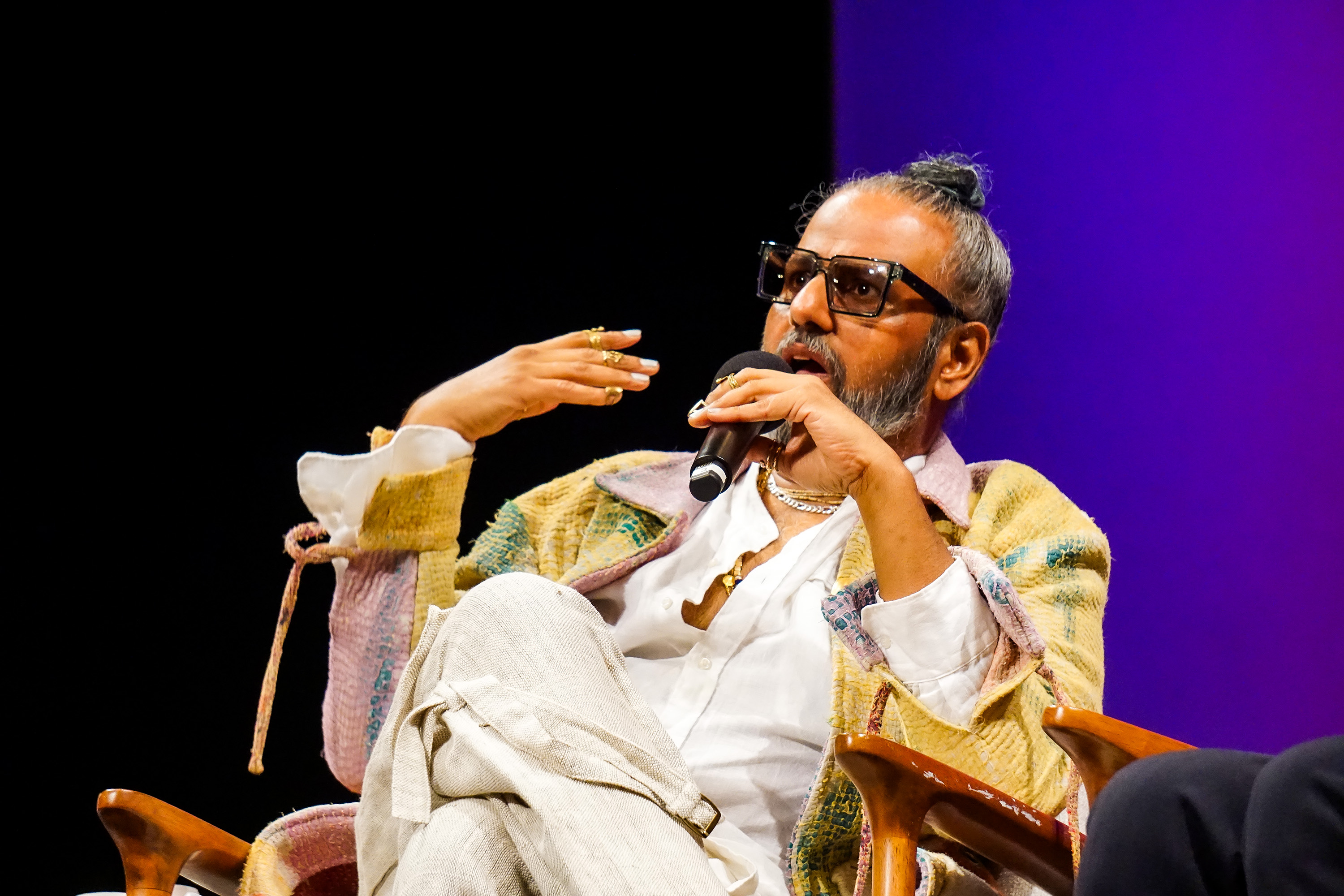 Ajay Shrivastav sitting on stage making a point while holding a microphone and wearing gold jewelry