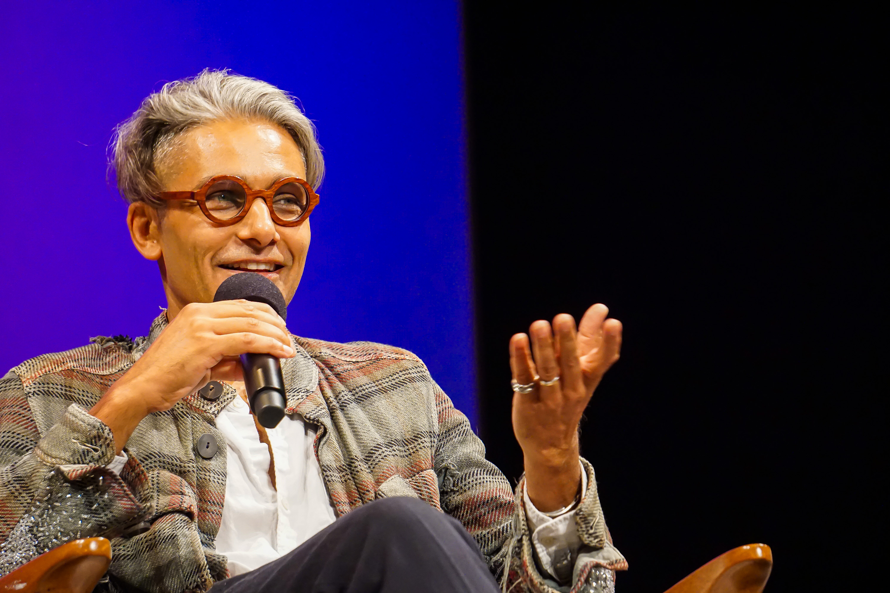 Sandy Dalal sitting and speaking into a microphone with his hand gesticulating while smiling