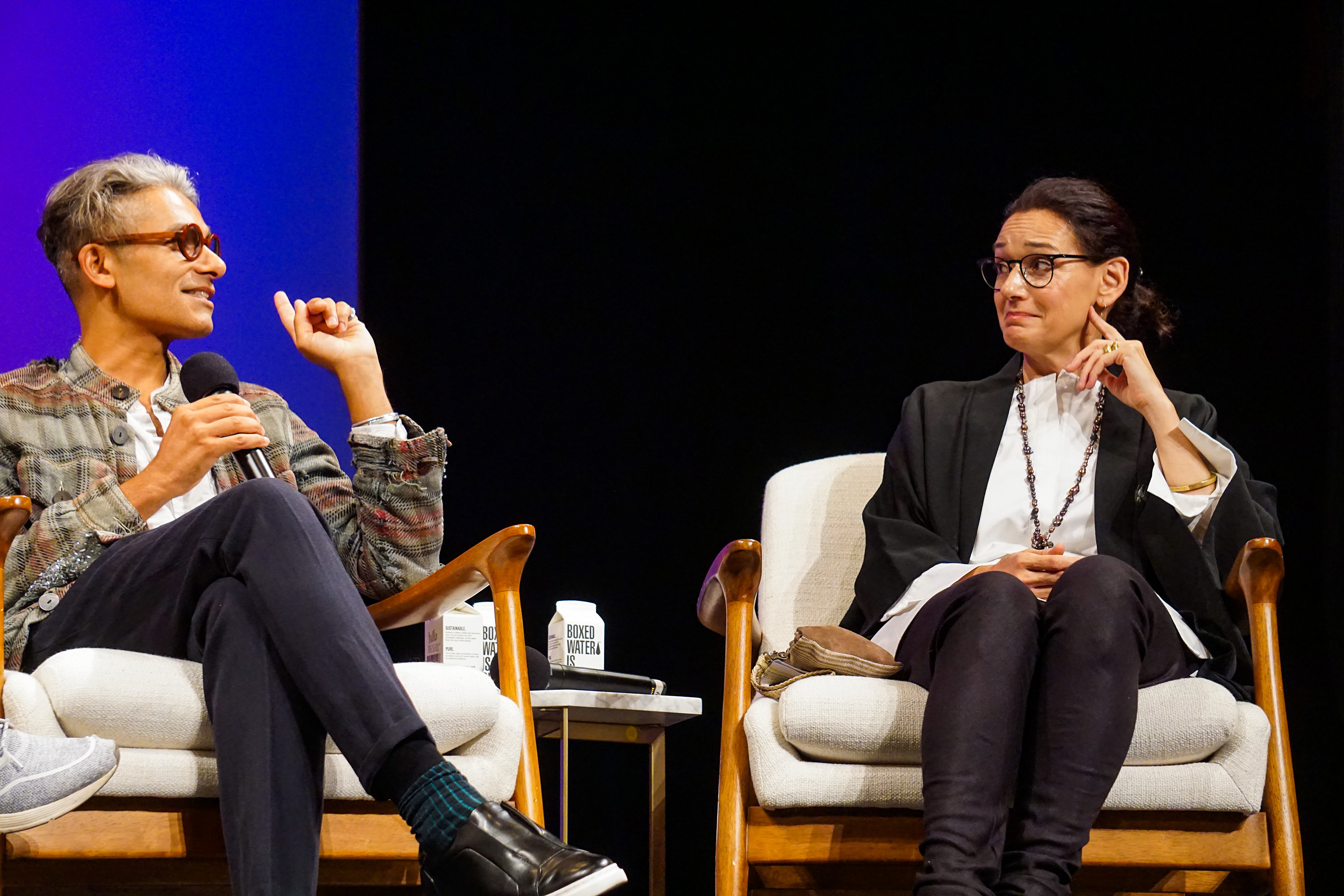 Sandy Dalal is making a point to Carmen Artigas who appears to be smirking, both sitting on stage