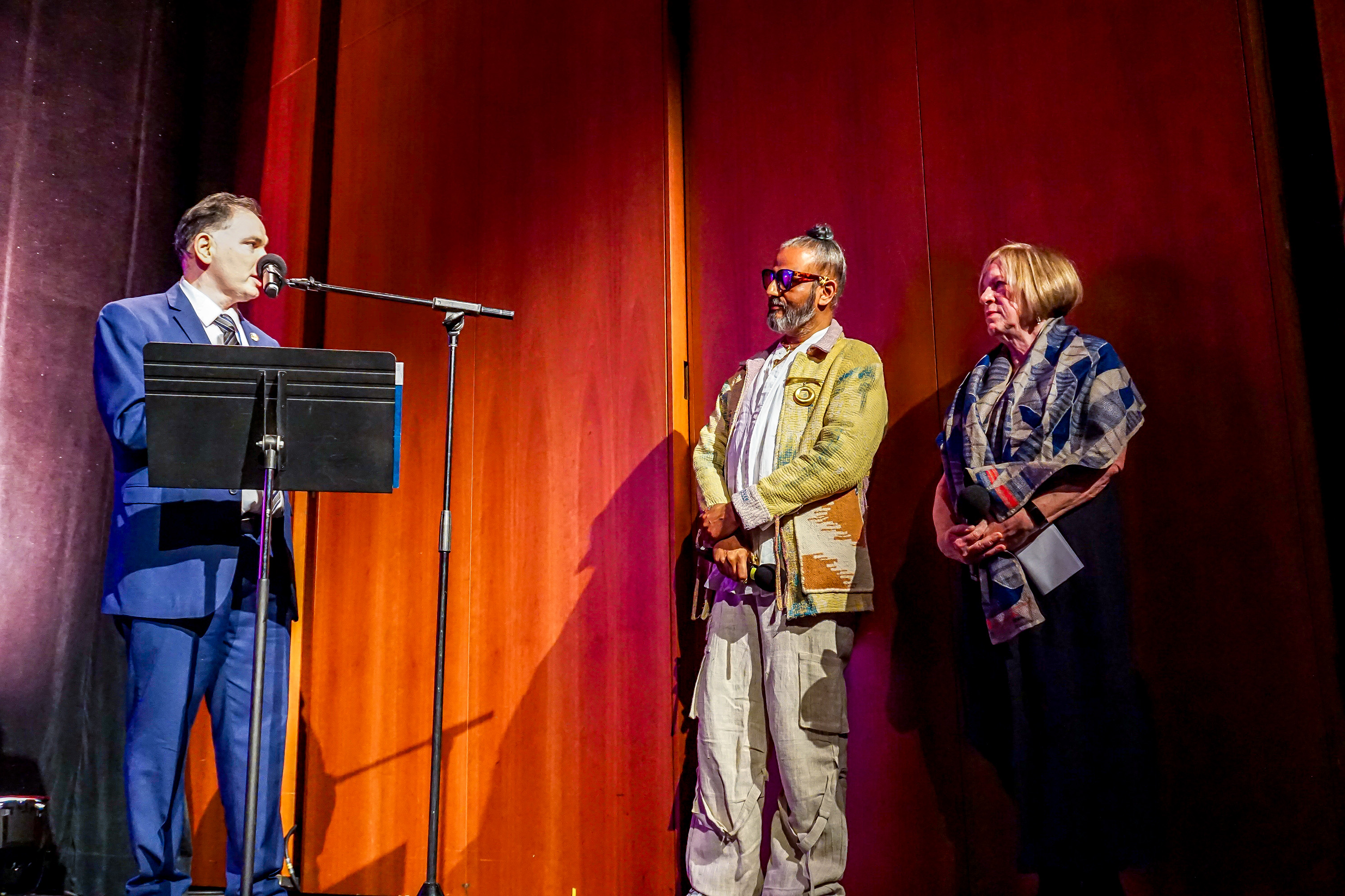 Ajay Shrivastav and Rachel Cooper standing next to each other on stage as a NY government rep speaks on podium