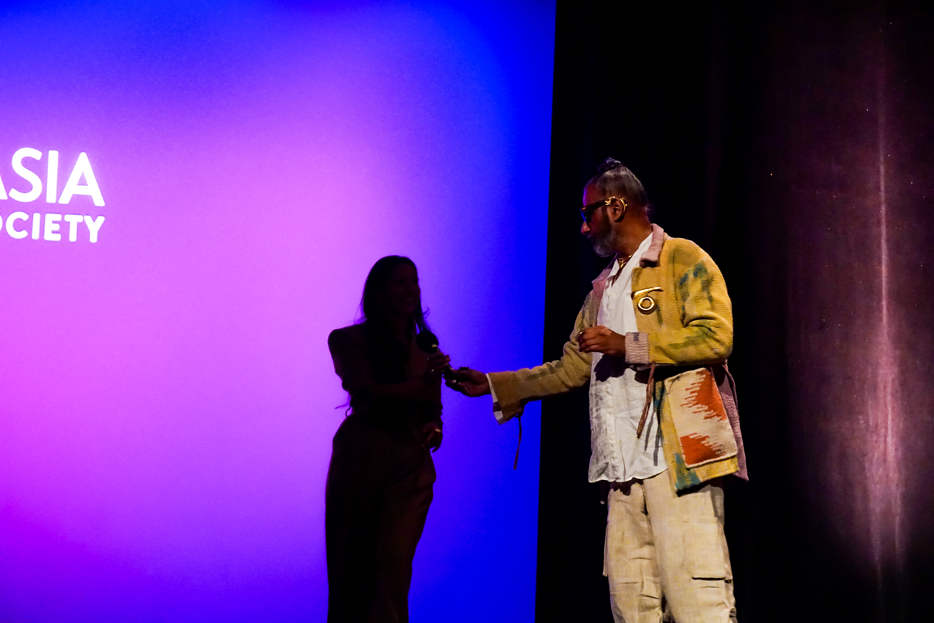 Ajay Shrivastav handing a silhouette of Poonam Thimmaiah the microphone on stage against a purple background