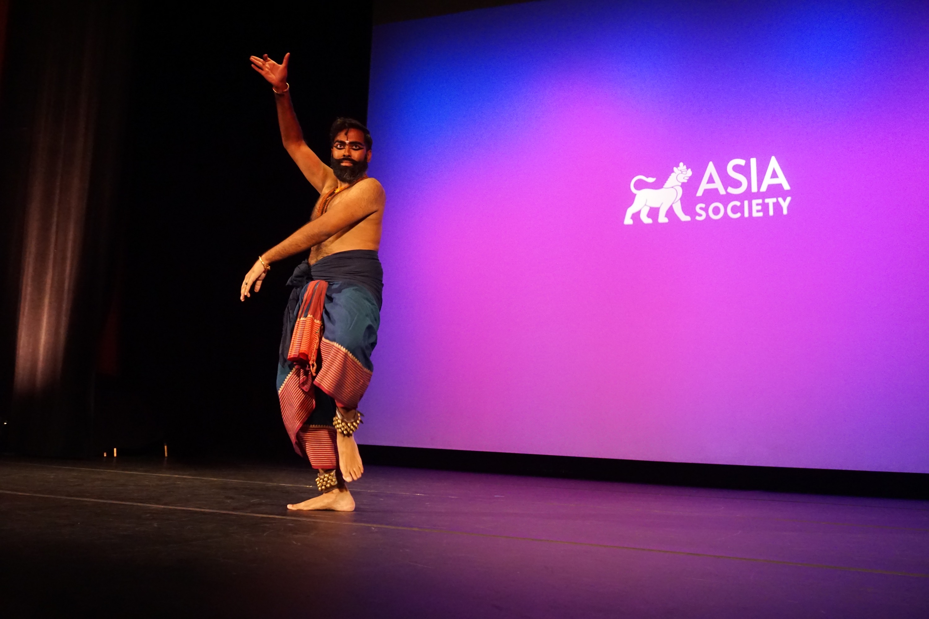 Jeeno Joseph dancing on stage without a shirt against a purple background with the Asia Society logo