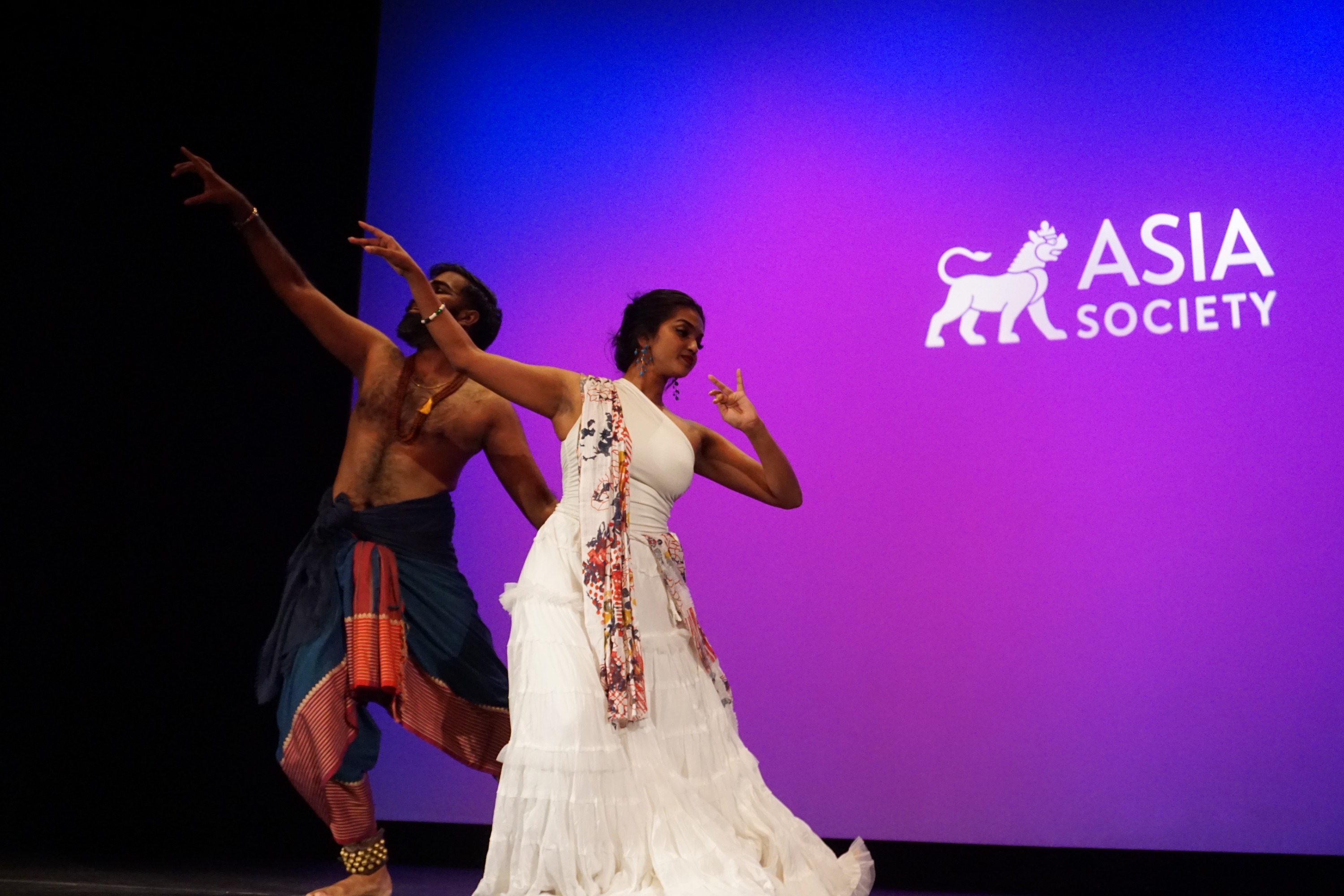 Jeeno Joseph and female model wearing Indian textiles and dancing with their hands in the air against a purple background on stage with the Asia Society logo