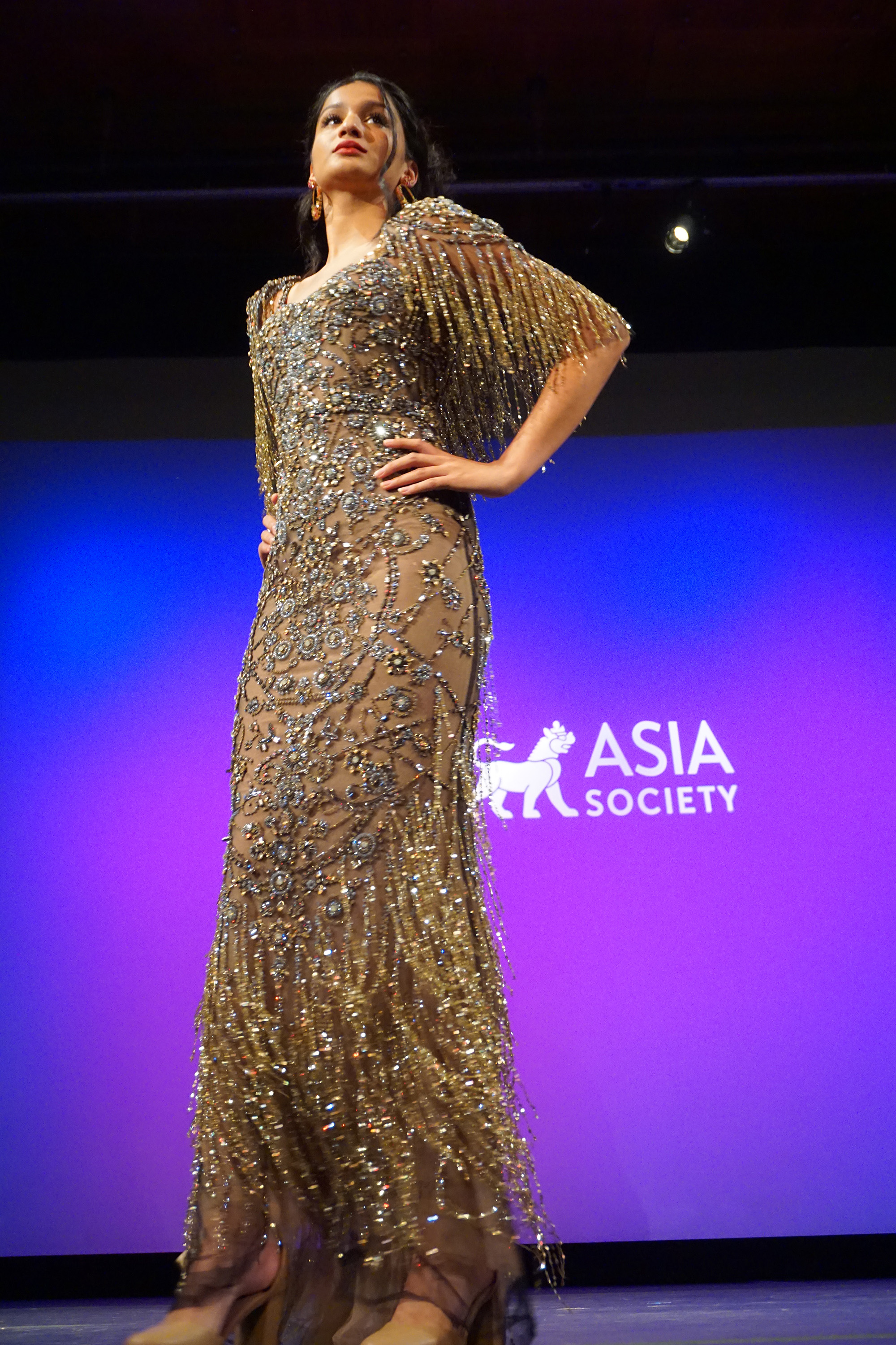 Female fashion model wearing a grey dress by Ajay Shrivastav posing on stage with a purple background with Asia Society logo