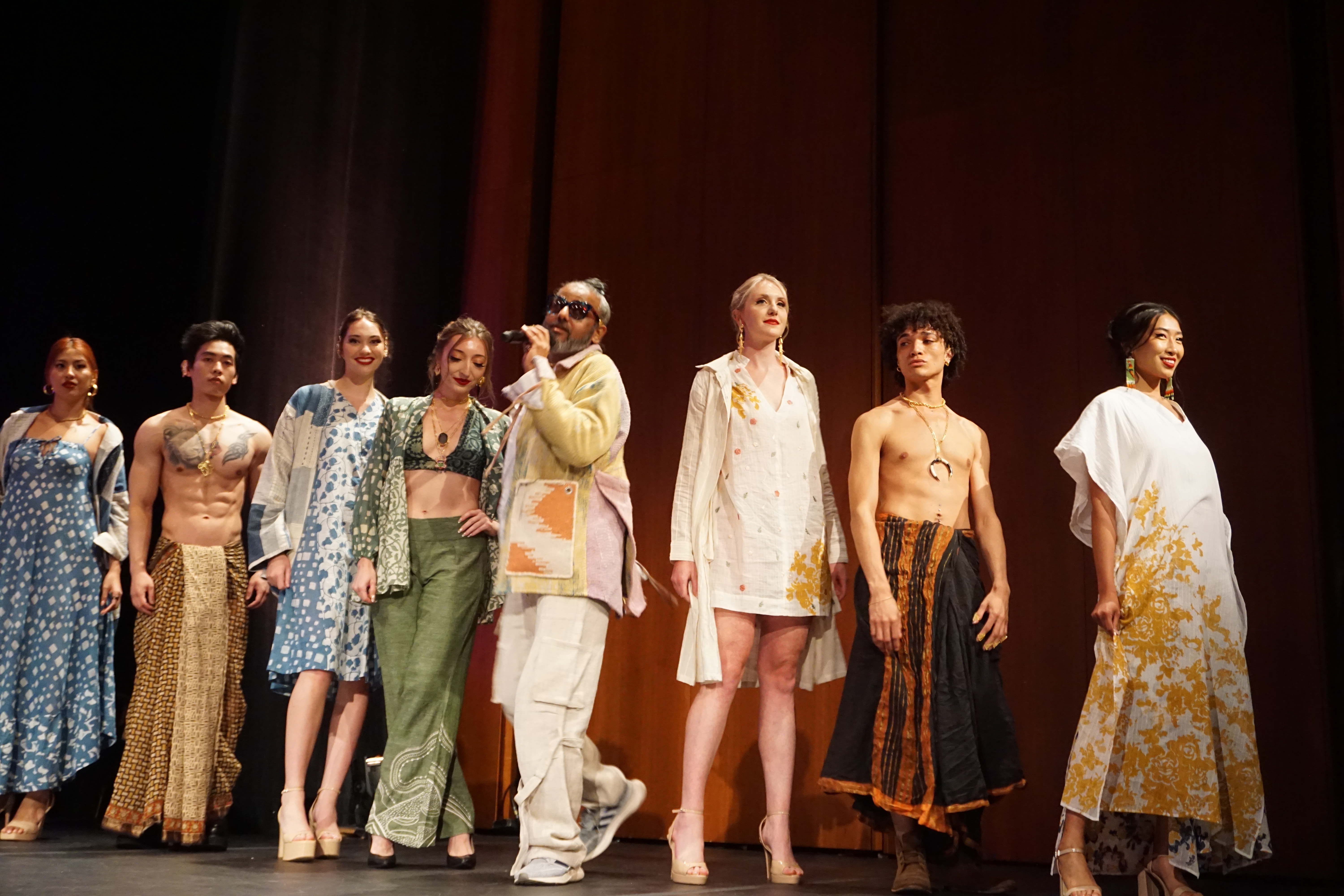 A line of models on stage with Ajay Shrivastav walking through the middle speaking on the microphone for the fashion show