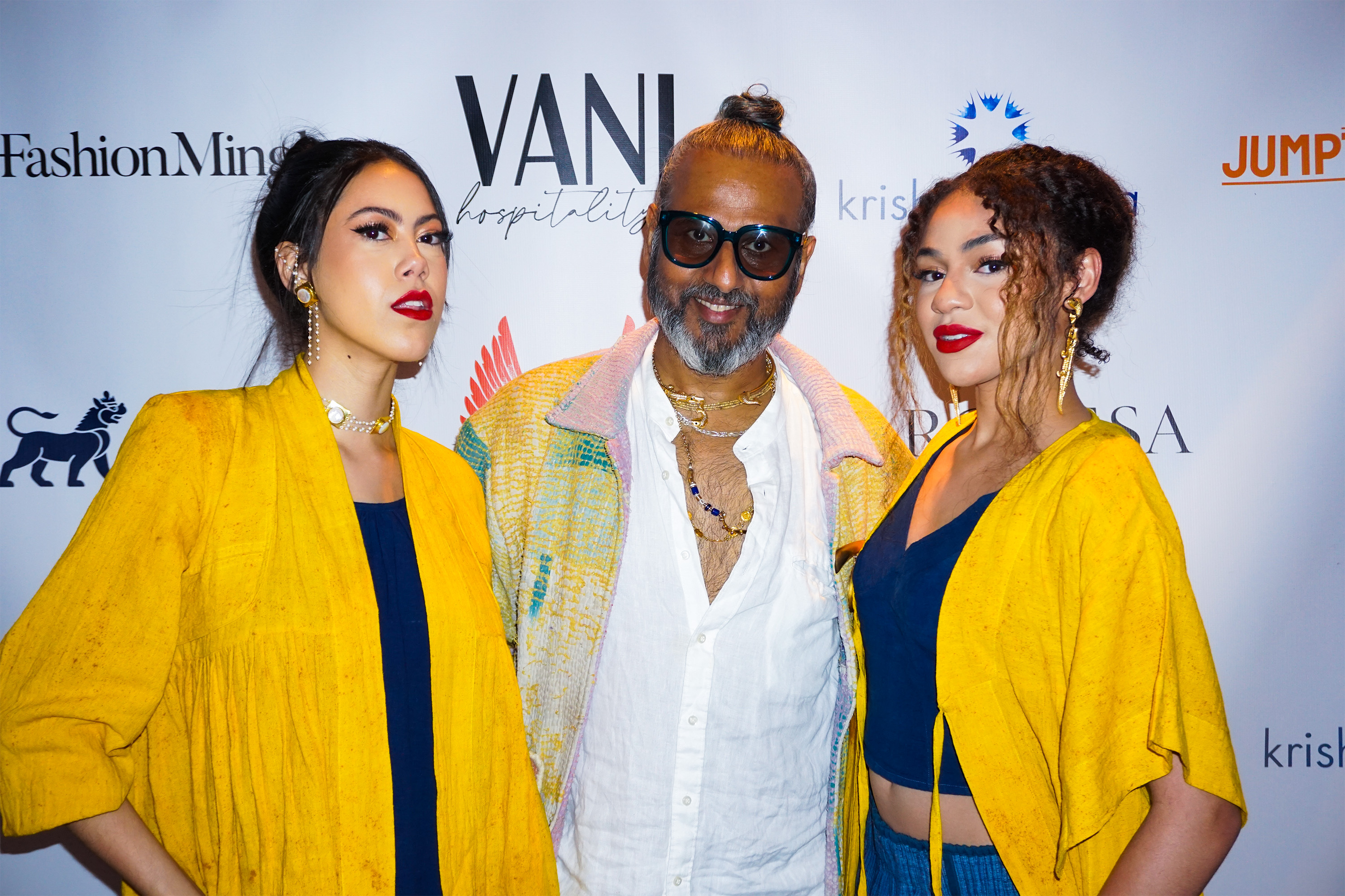 Ajay Shrivastav standing in between two female models posing wearing bright yellow clothing  in front of a step and repeat banner