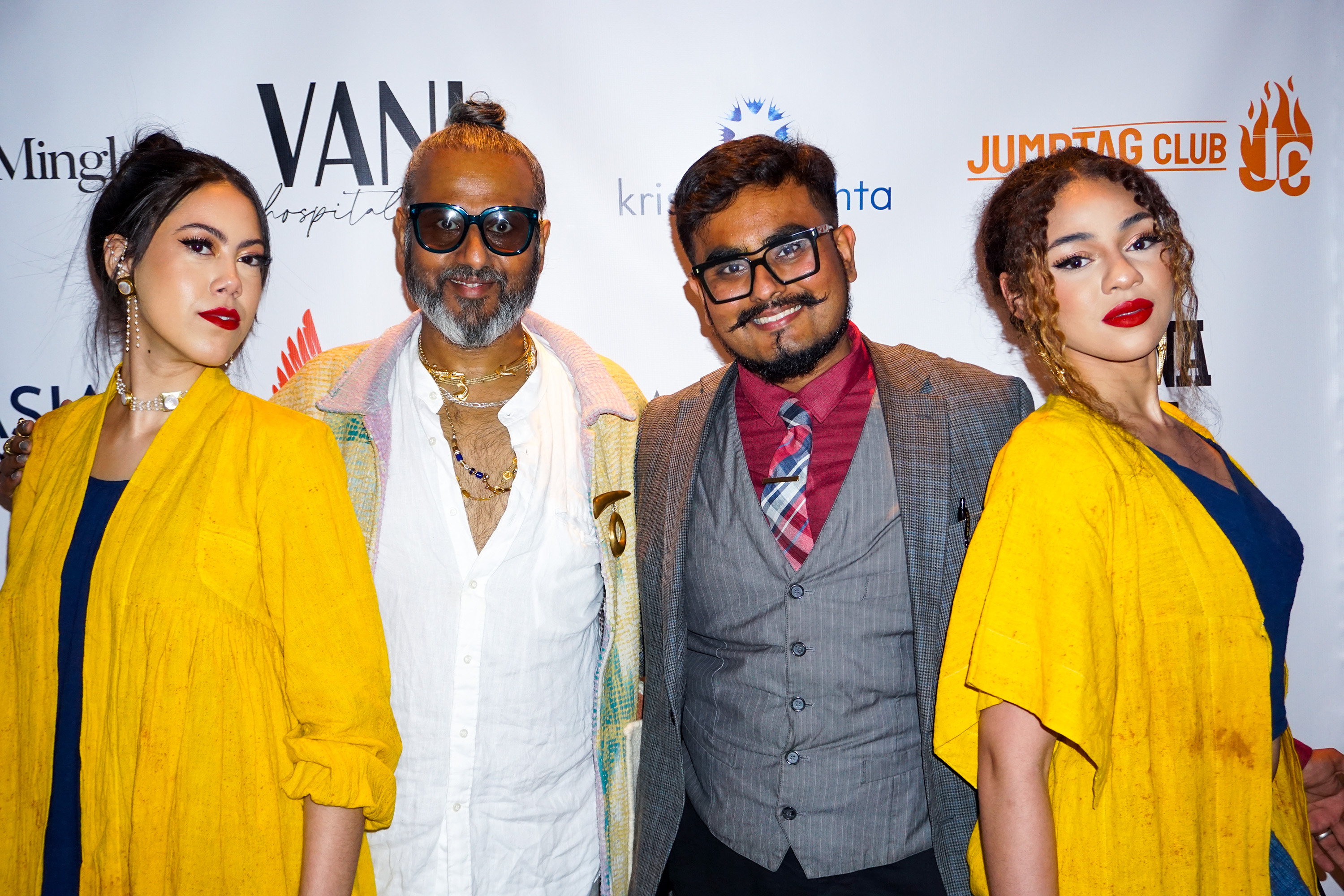 Ajay Shrivastav and his son standing in between two female models wearing bright yellow clothing  in front of a step and repeat banner