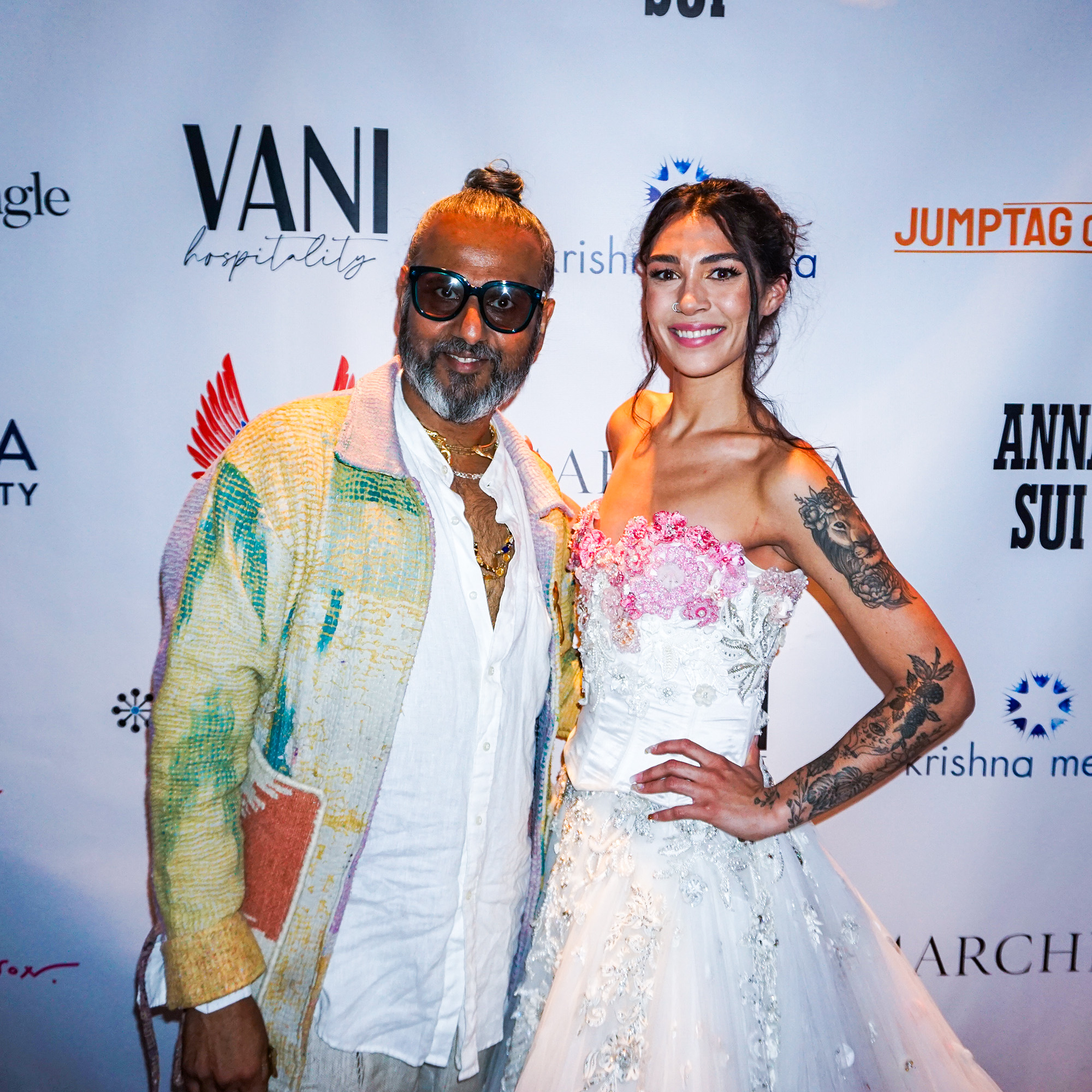 Ajay Shrivastav and a female model wearing a white dress in front of a step and repeat banner