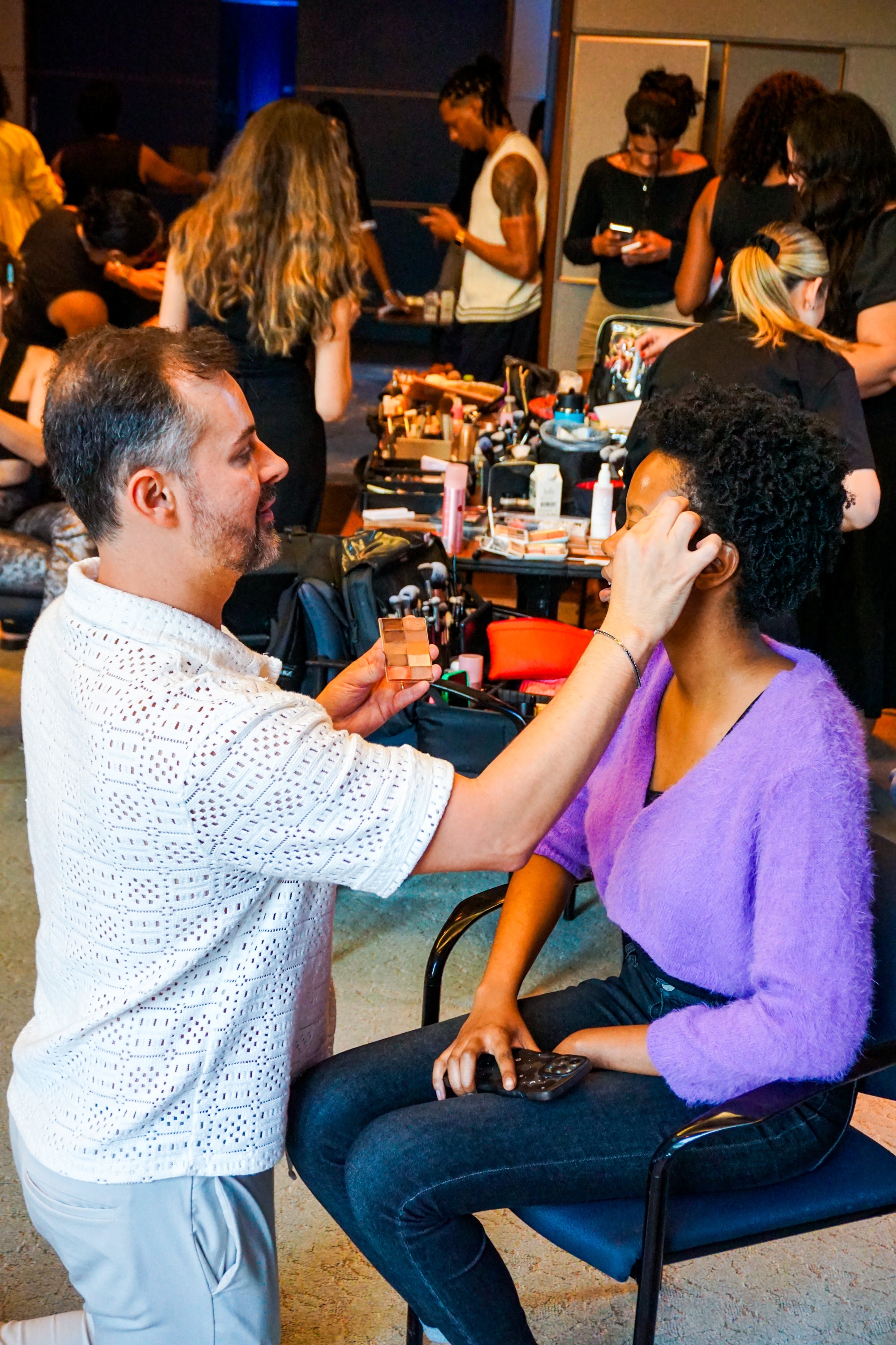 A male make up artist kneeling as he applies make up to a female model sitting in a chair with a busy background backstage