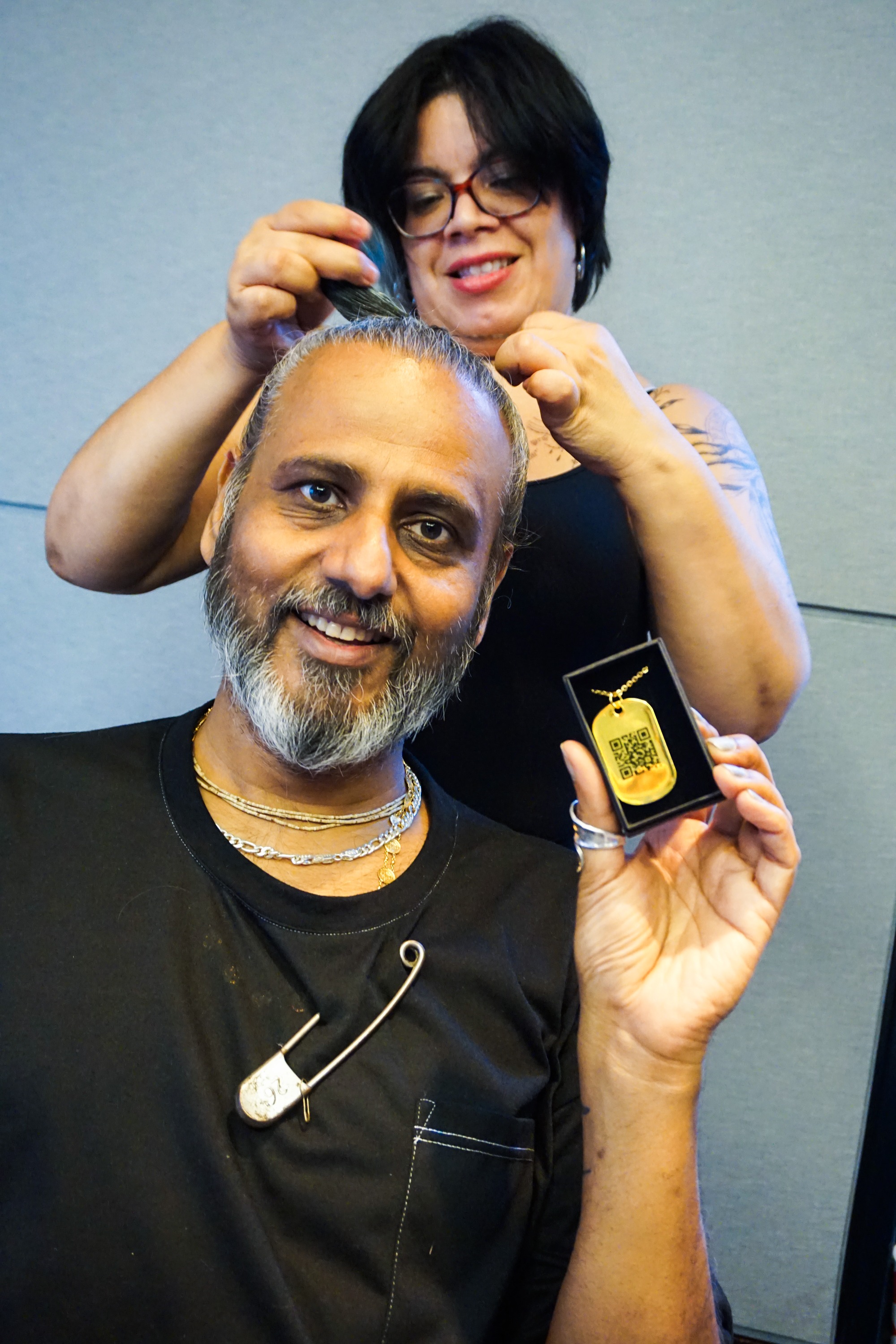 Hair stylist doing Ajay Shrivastav's hair while he holds a golden jumptag dog tag in its box to the camera smiling