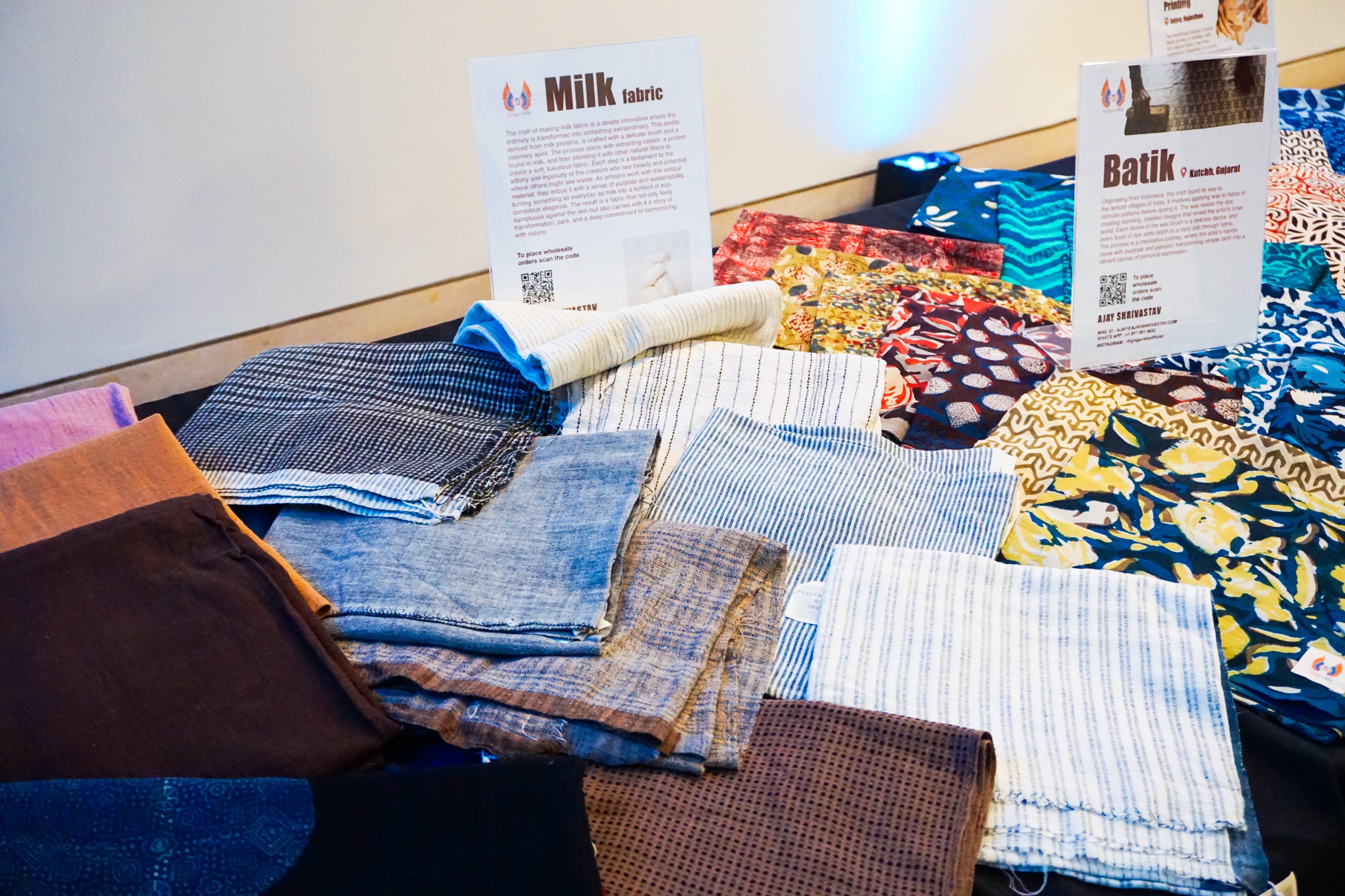 a variety of patterned textiles made by Ajay Shrivastav laid out on a table with signs indicating their style and materials