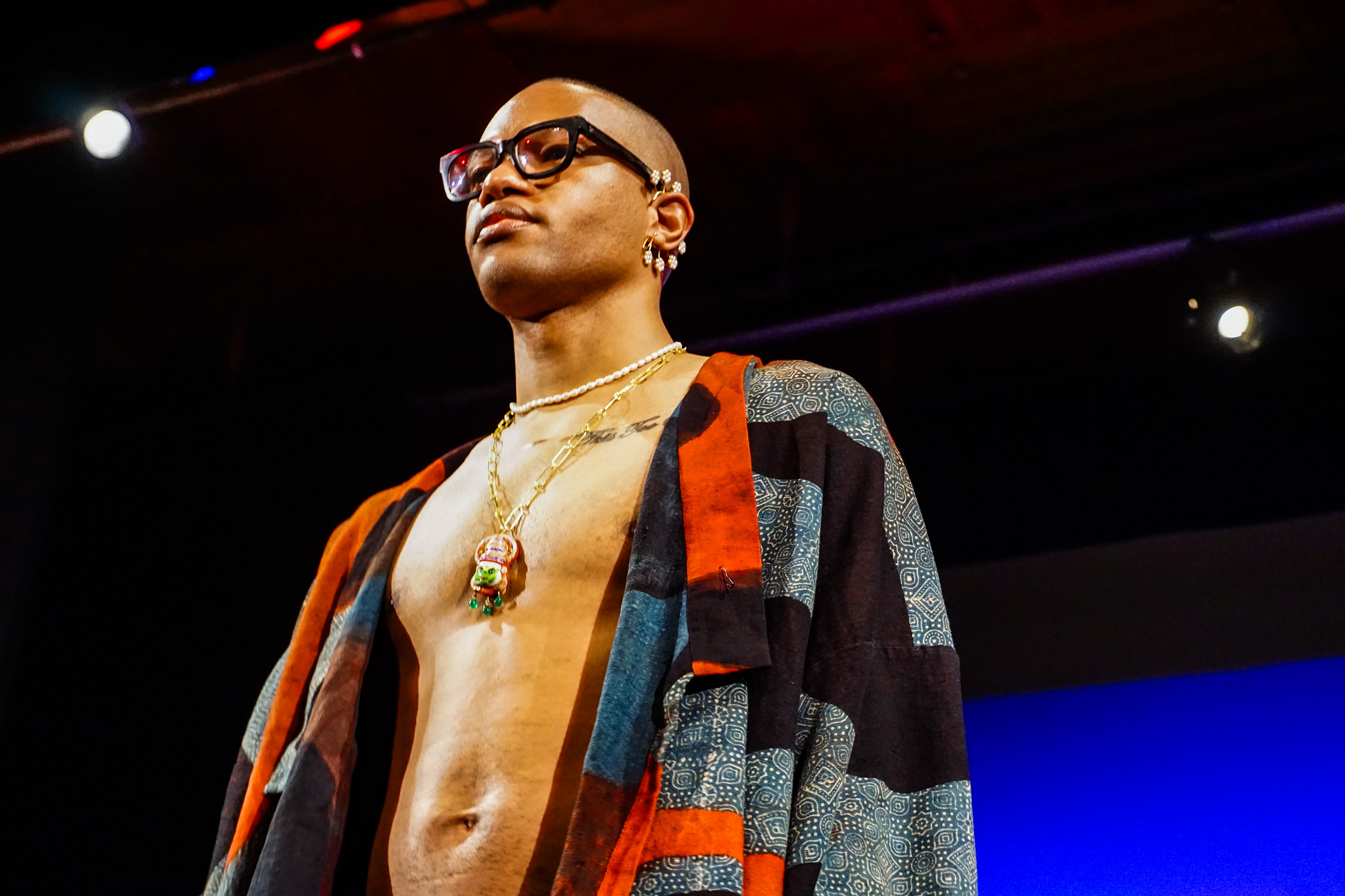 Close up of African American male model with glasses, chest exposed and a patterned red, black and grey jacket on runway