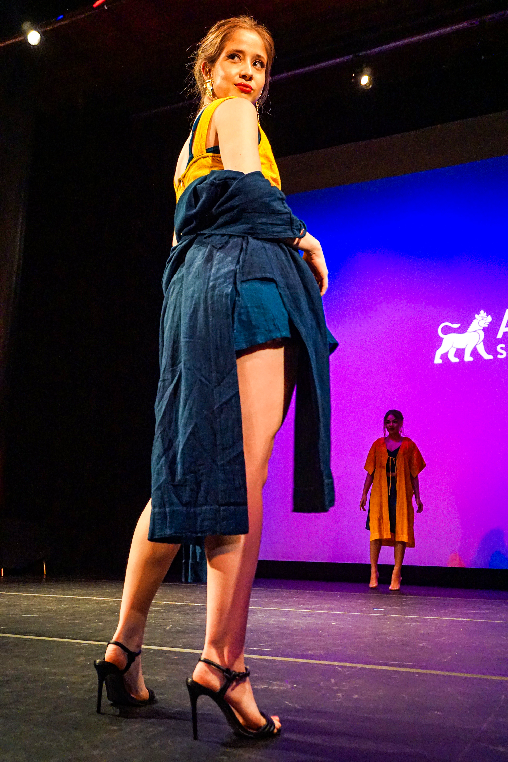 Female model with blue and yellow fashion attire posing with her back turned and head twisted on the runway to the crowd