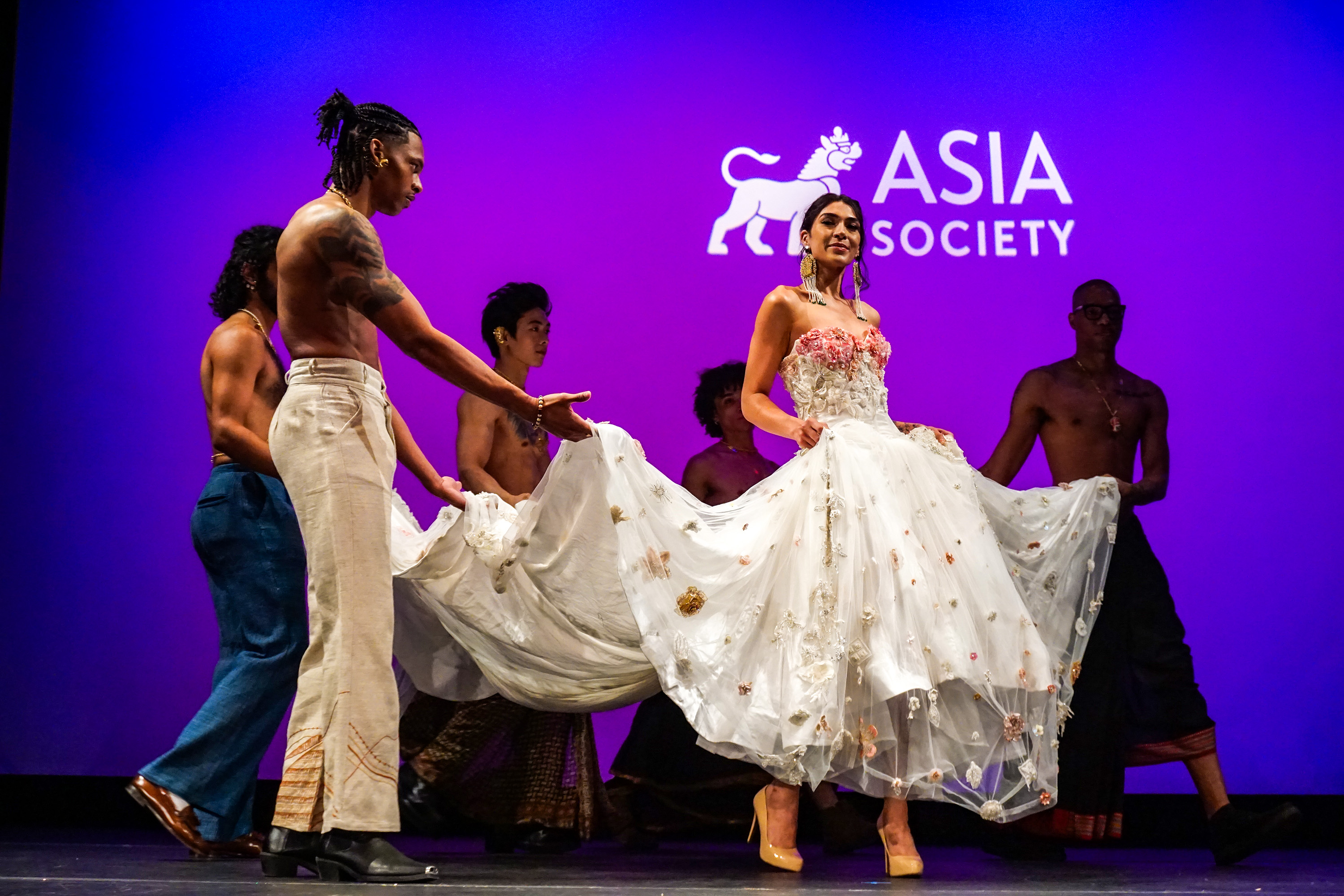 Female model with long white dress walking on runway stage with group of male models carrying bottom of her dress