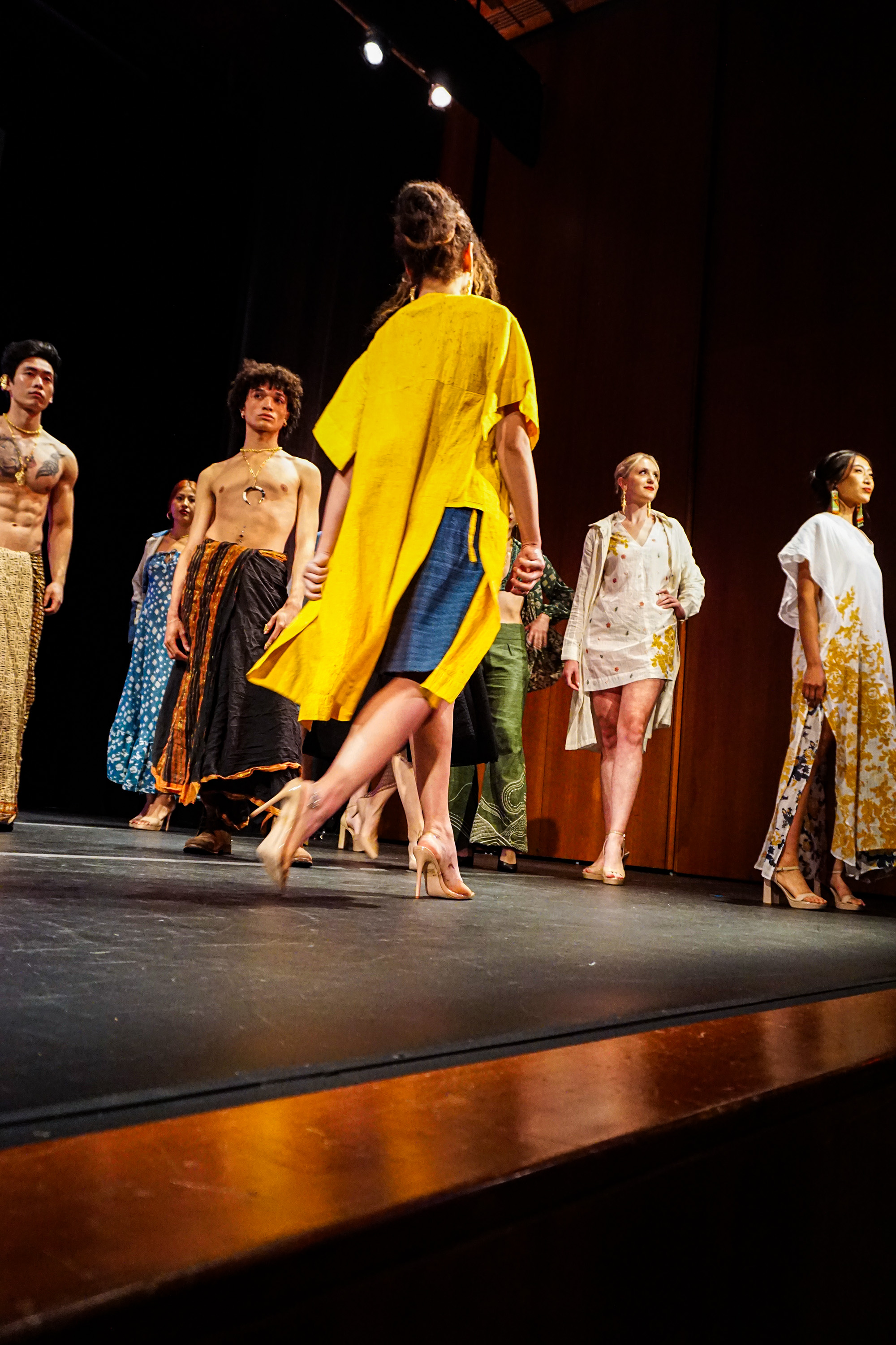 Female model in bright yellow fashion attire walking off runway with other models in the background