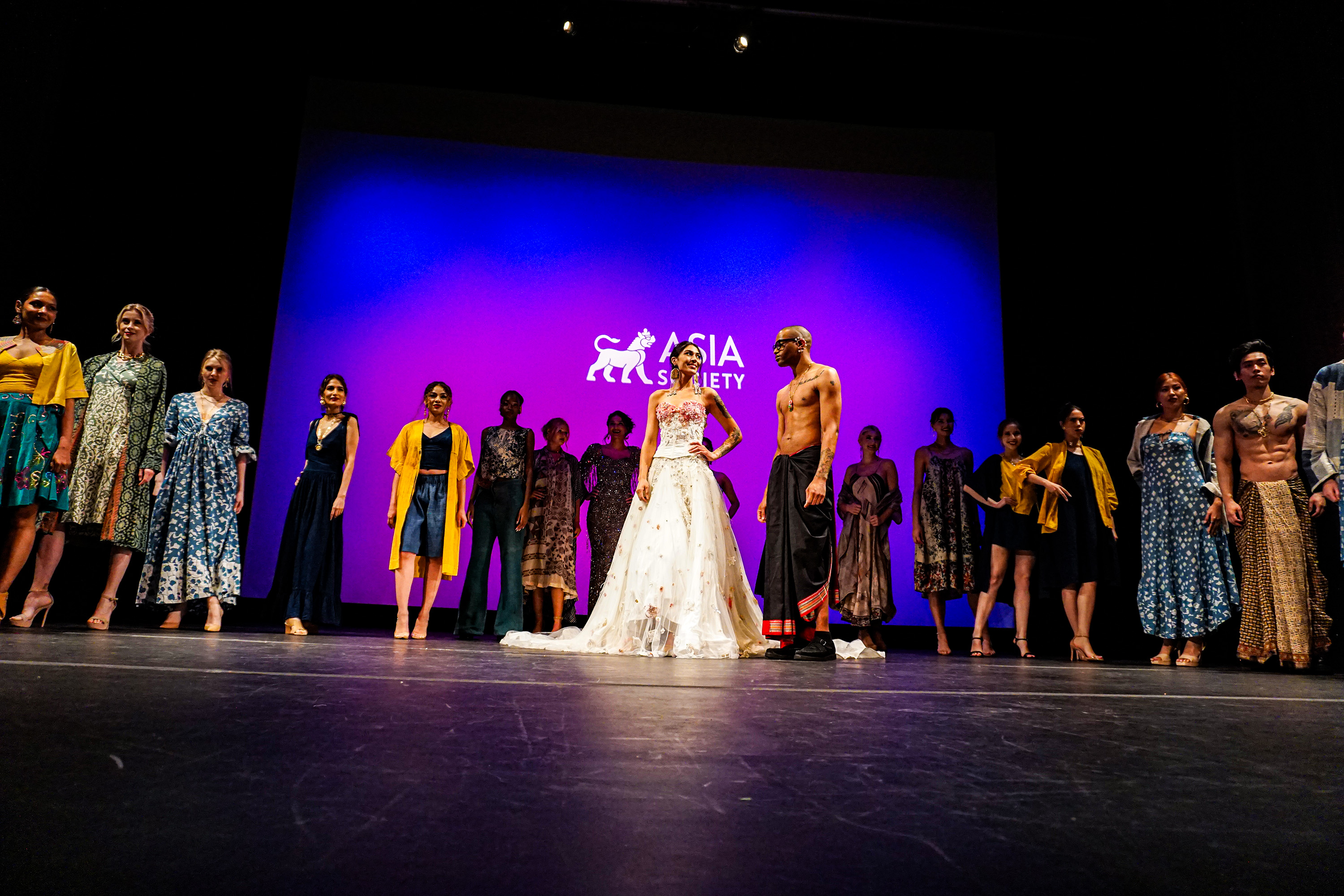 A crowd of models lined up on the runway stage with a woman with a long white dress in the middle