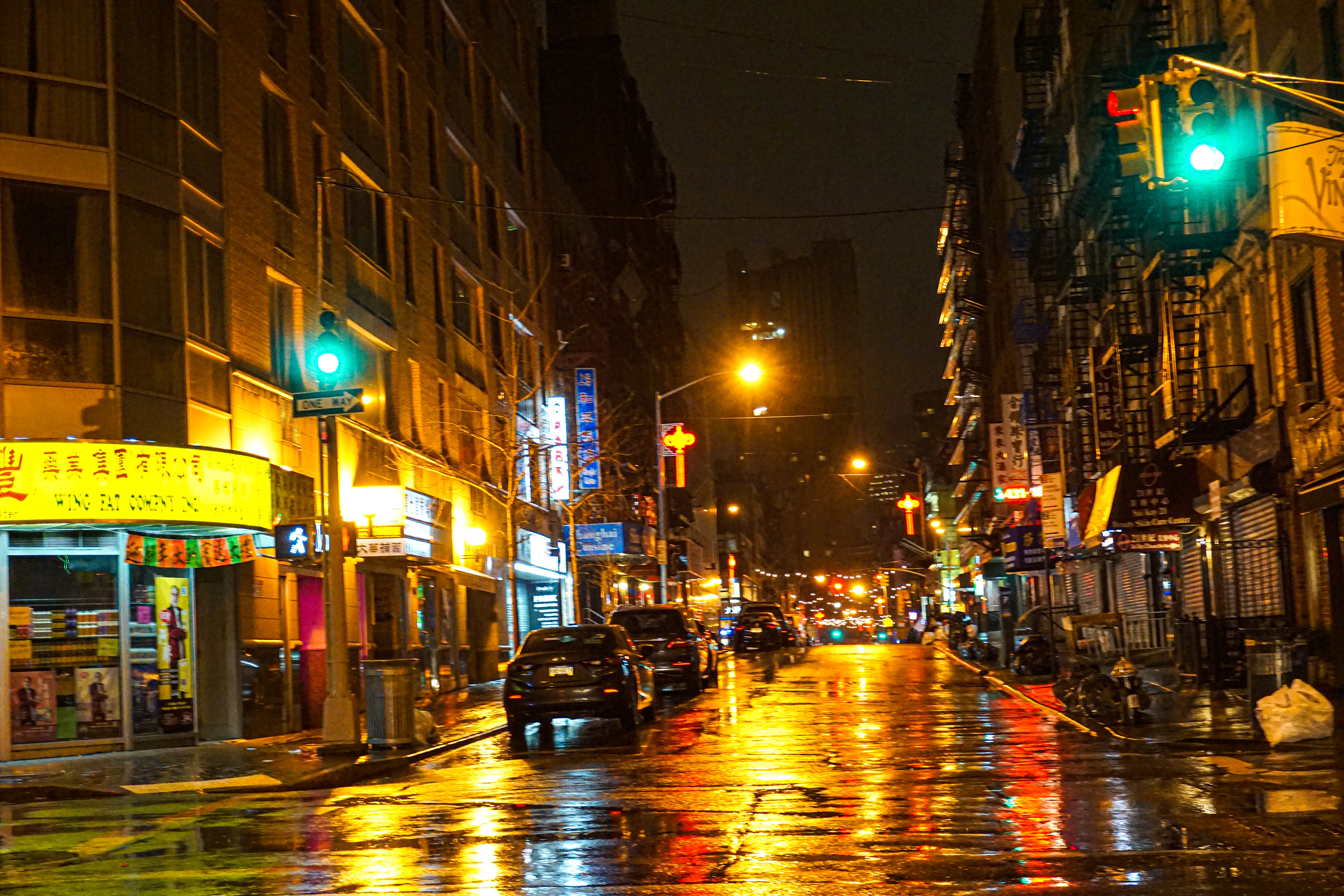 A shiny, reflective street in Chinatown New York City night time after it rained, with glistening parked cars and lights