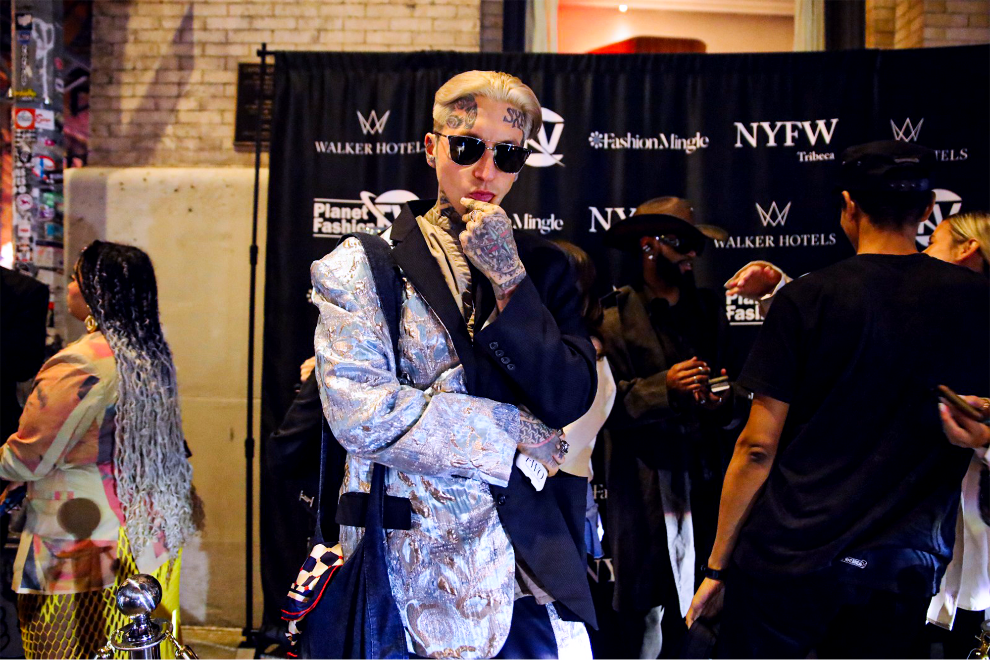 Clavish posing with his hands on his chin wearing a half silver half black blazer in front of step and repeat banner 