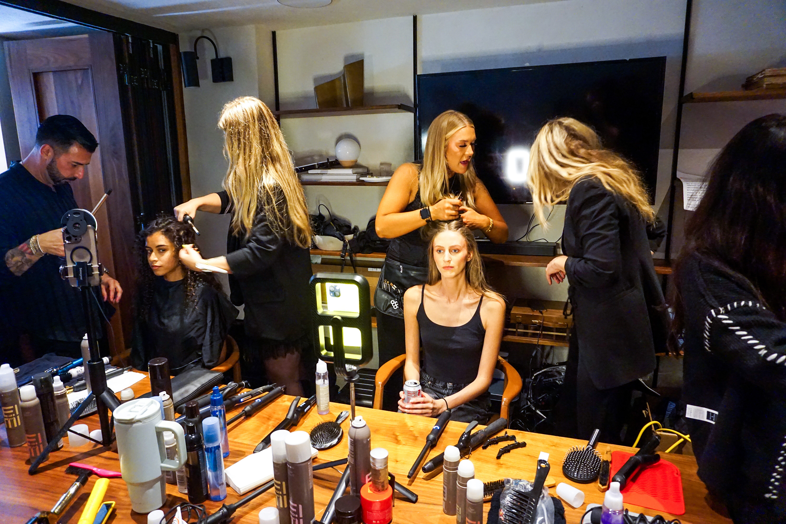 A busy room of hair and make up artists working with models with supplies all over a table