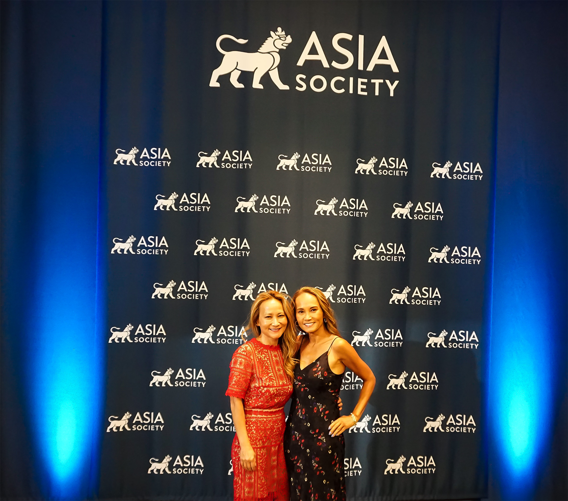 Julie Tran and friend posing for camera in front of step and repeat banner with Asia Society logos and blue lights