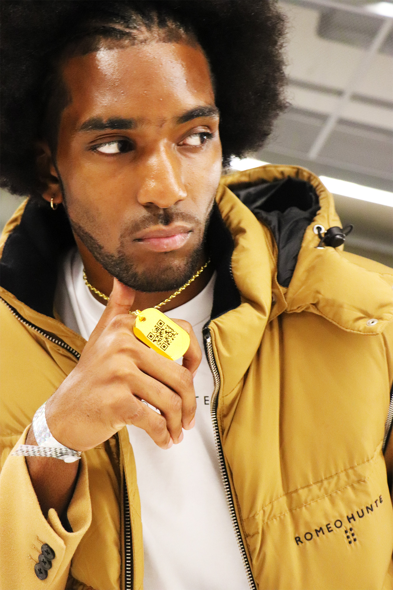 Close up of African American male model wearing Romeo Hunte jacket and holding out golden jumptag dog tag with its QR visible