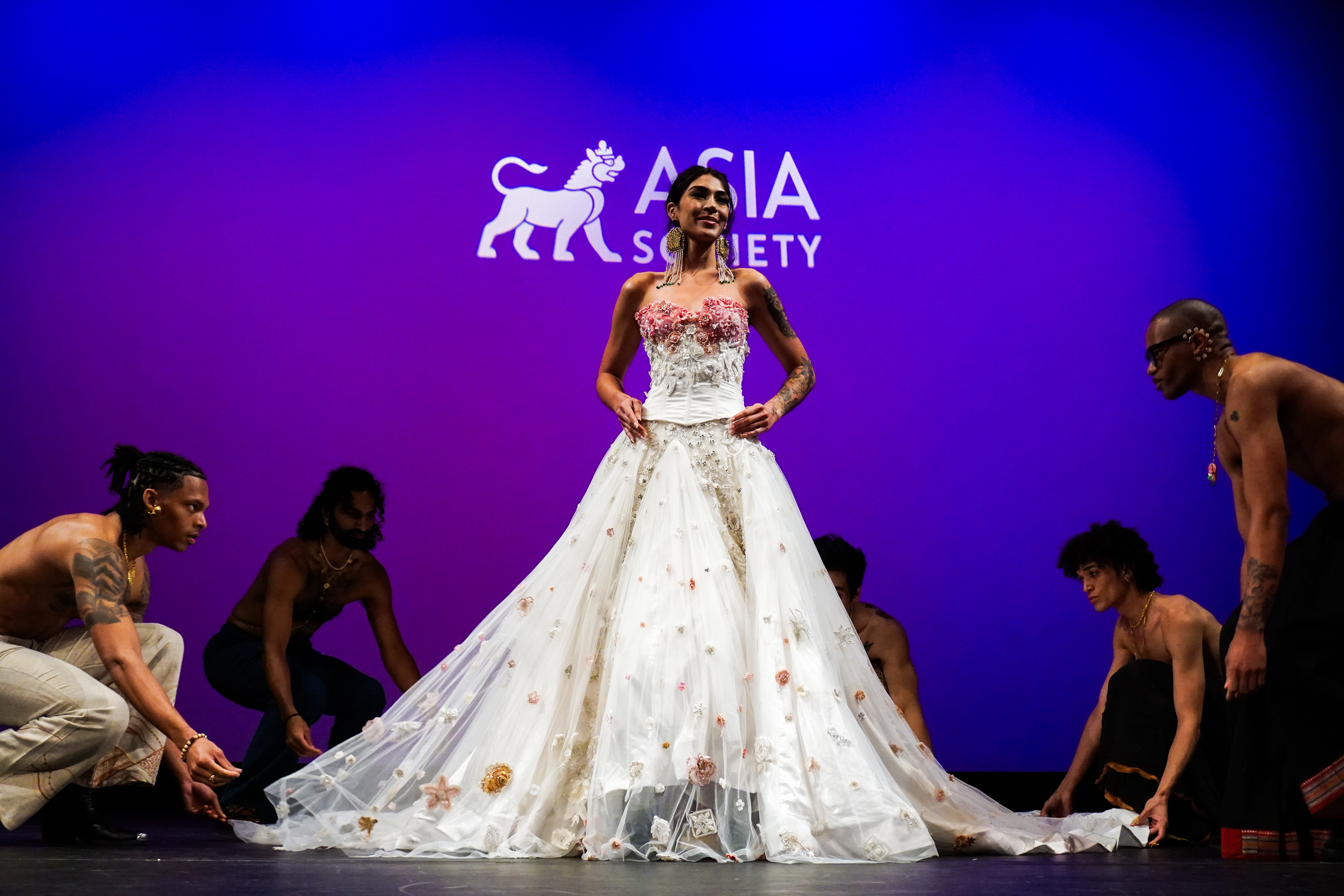 Model on Asia Society New York stage wearing long white dress surrounded by male models grabbing bottom of her dress