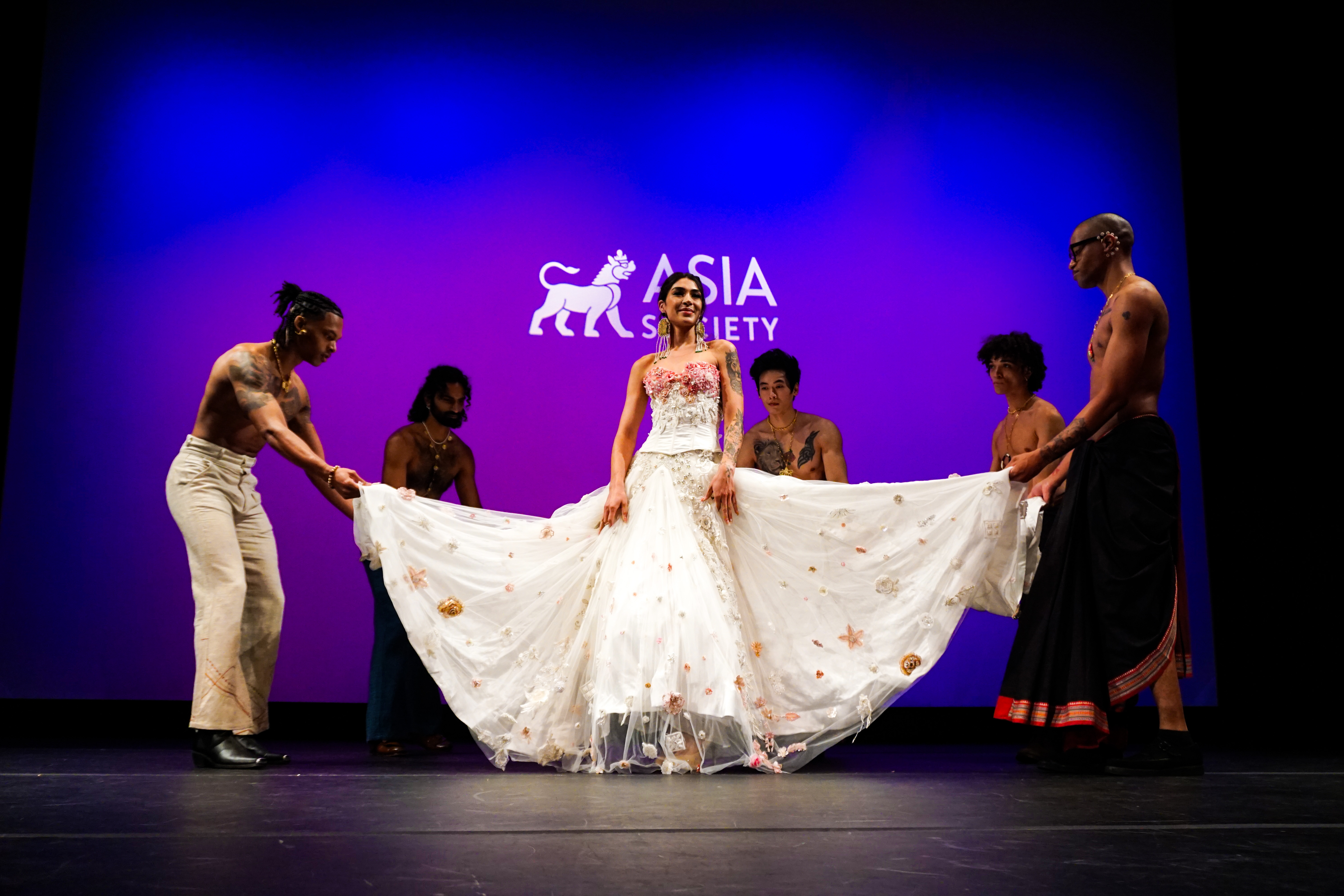 Model on Asia Society New York stage wearing long white dress surrounded by male models lifting up the bottom of her dress