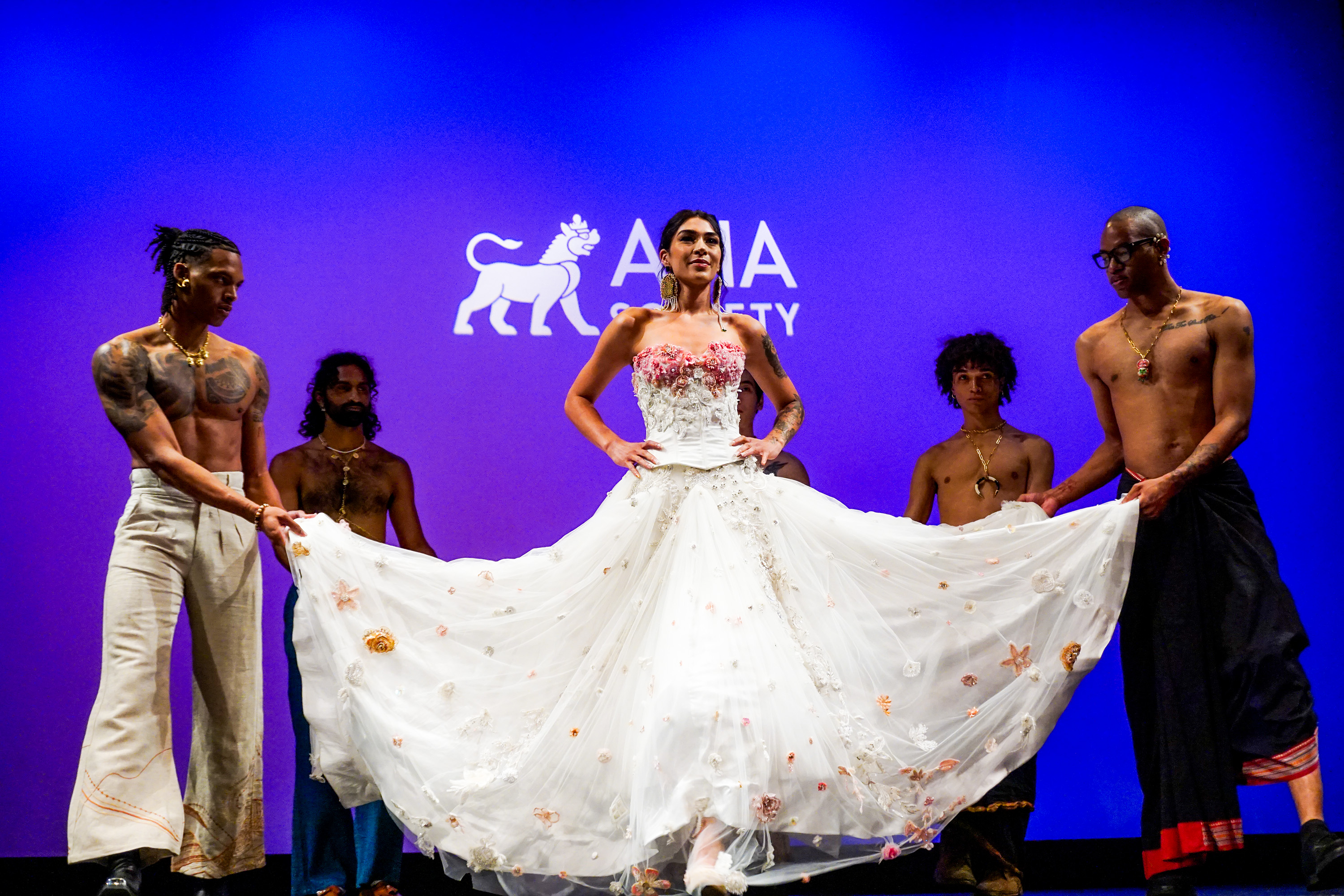 Model on Asia Society New York stage wearing long white dress surrounded by male models holding up the bottom of her dress