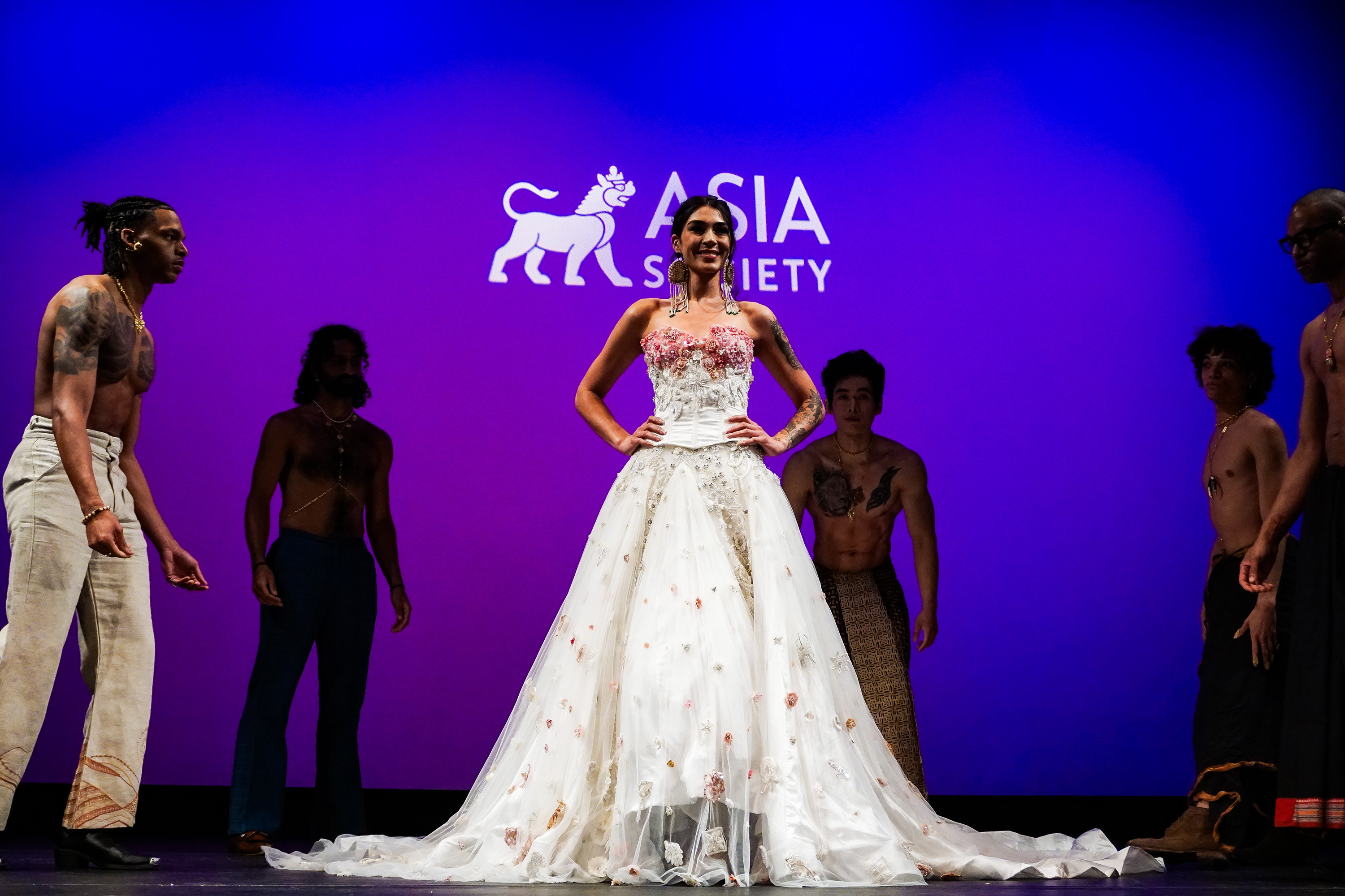 Model on Asia Society New York stage wearing long white dress surrounded by standing male models