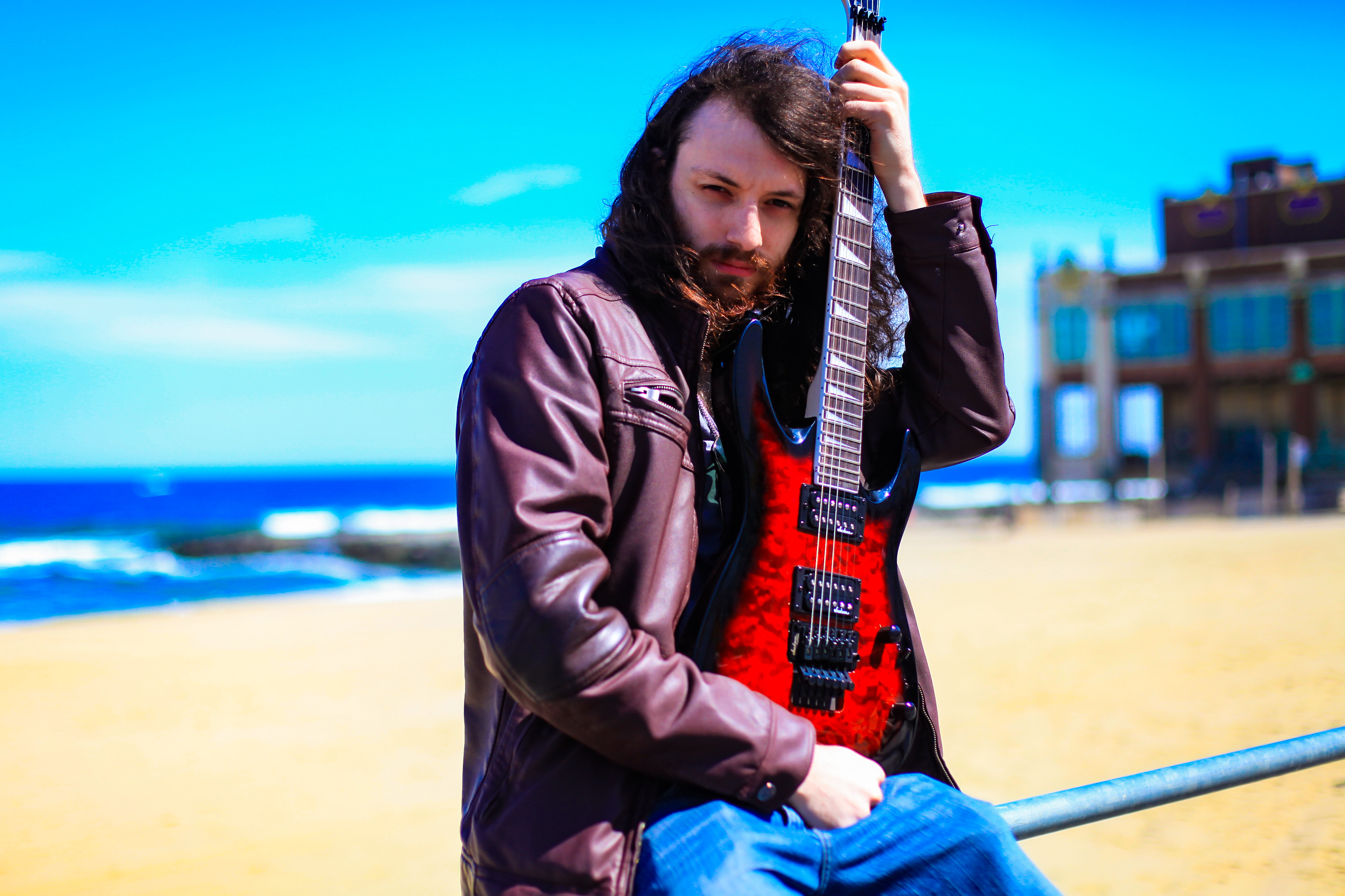 Rito holding a guitar on his lap to his face with a leather jacket sitting on a bannister with the beach behind him