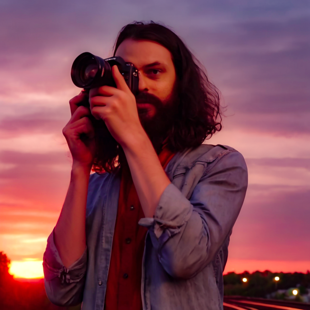 Rito holding a camera to his eye focusing on a shot with a sunset background