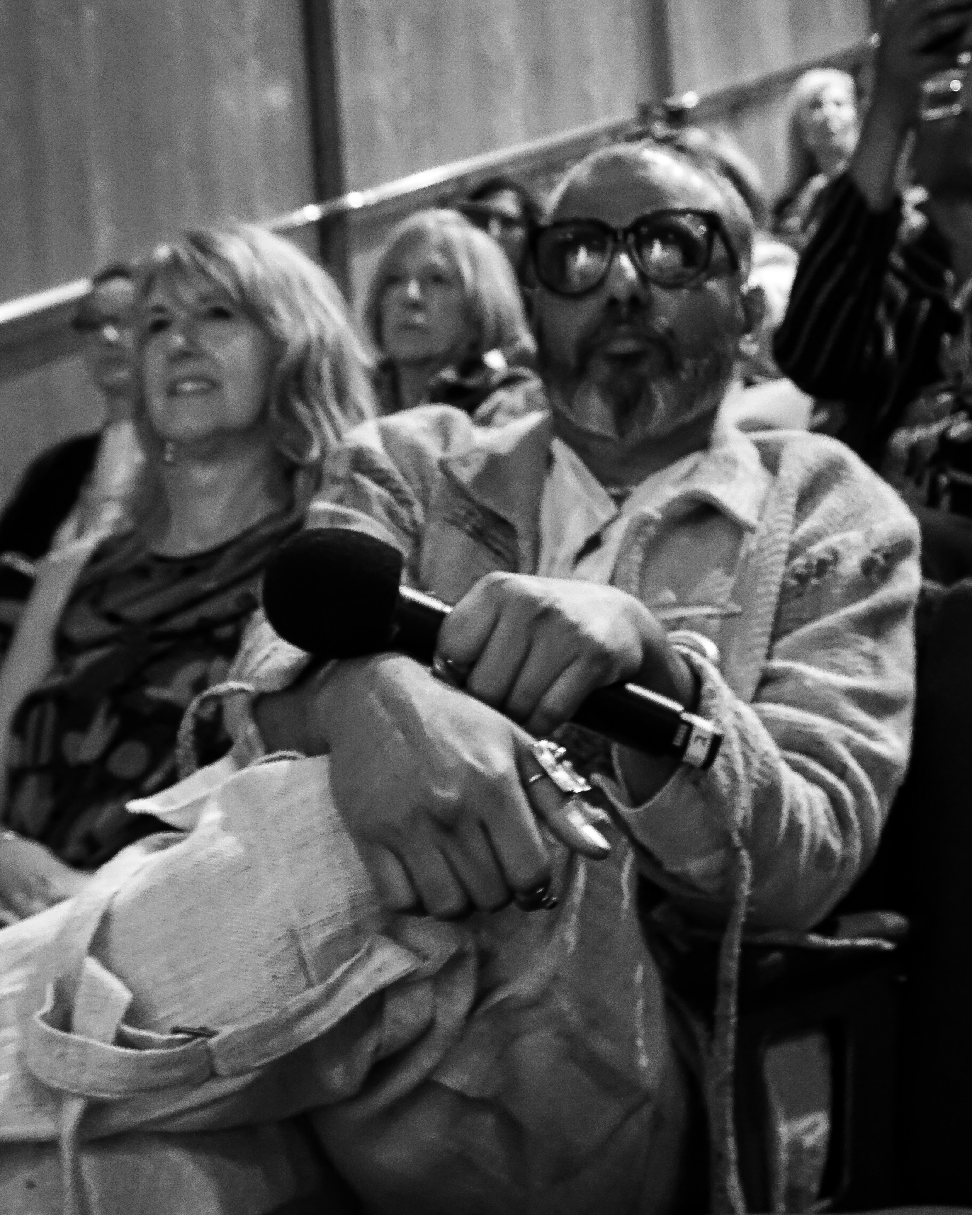 Ajay Shrivastav in black and white sitting in the audience watching a show with his legs crossed and a microphone in his hand