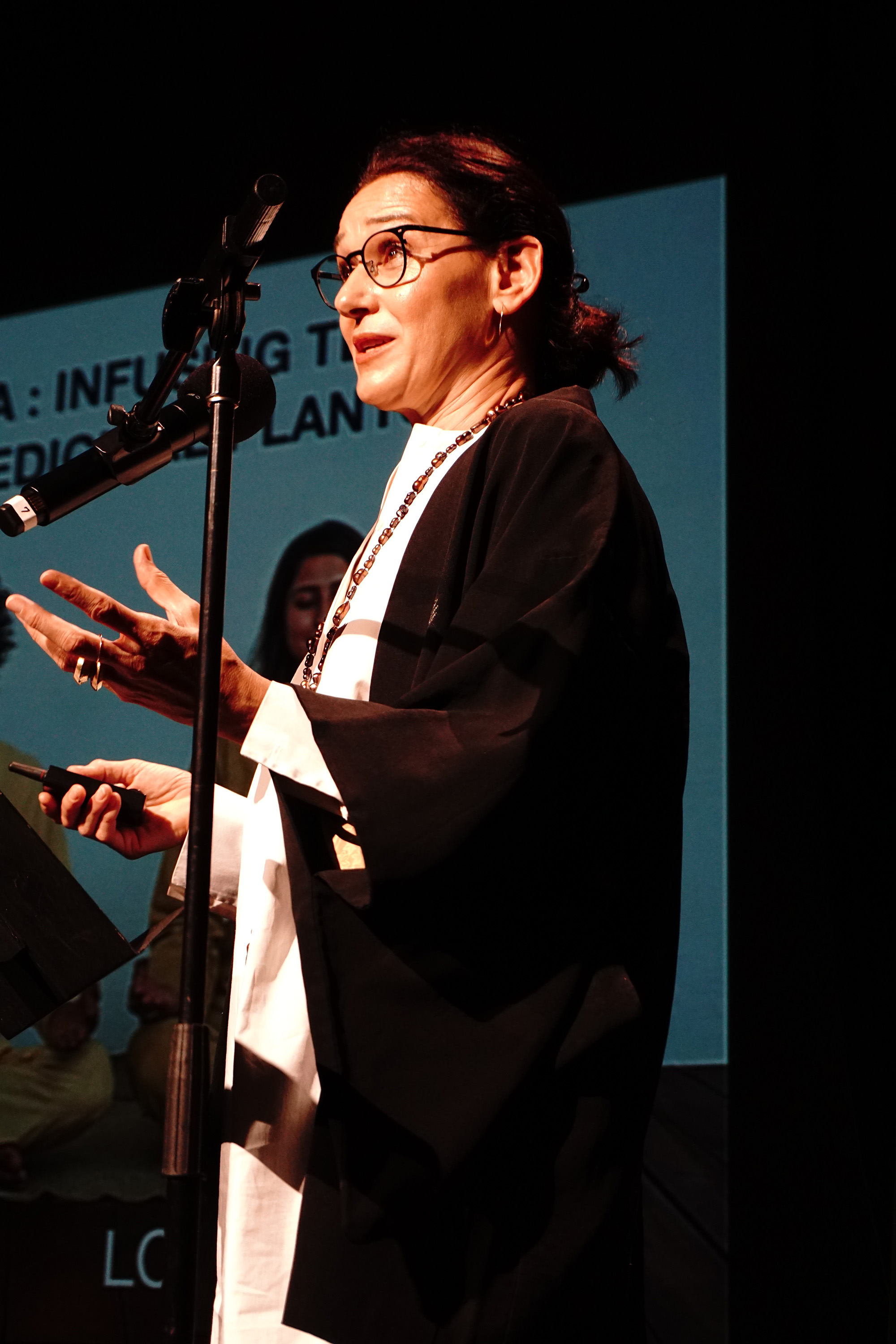 Carmen Artigas speaking on stage into a microphone in front of a slideshow projection screen