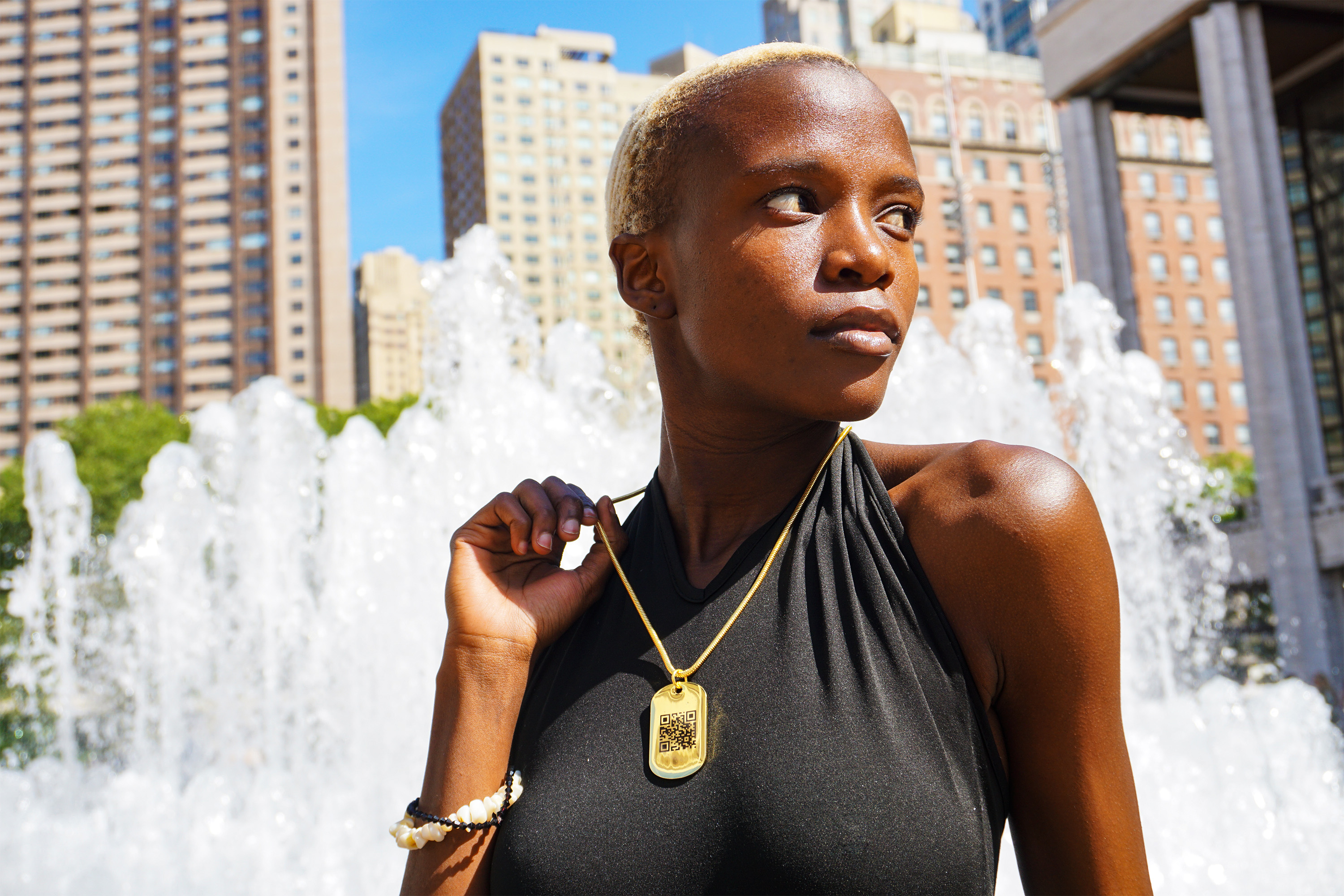 Female model sitting in front of a large New York fountain bustling water wearing a black shirt and a golden jumptag necklace