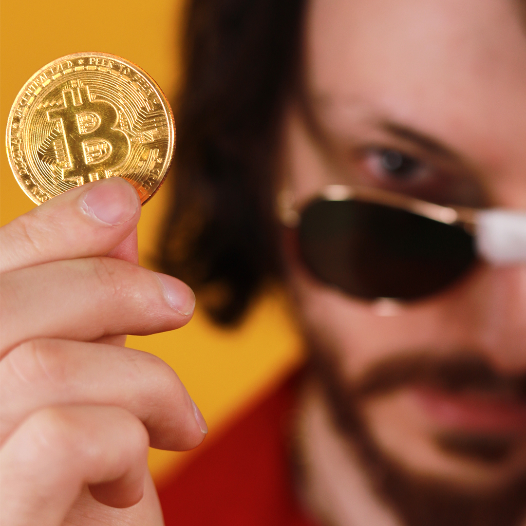 Close up of Rito holding a Bitcoin coin with half his face showing wearing sunglasses with tape in the middle